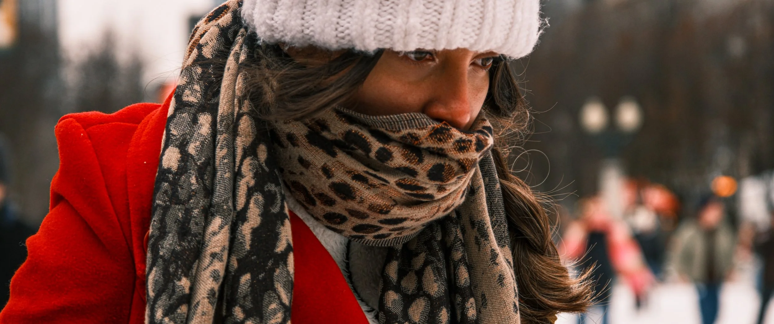 Close-up of a woman wearing a white knit hat, leopard print scarf, and red coat outdoor in cold weather, with people in the background.
