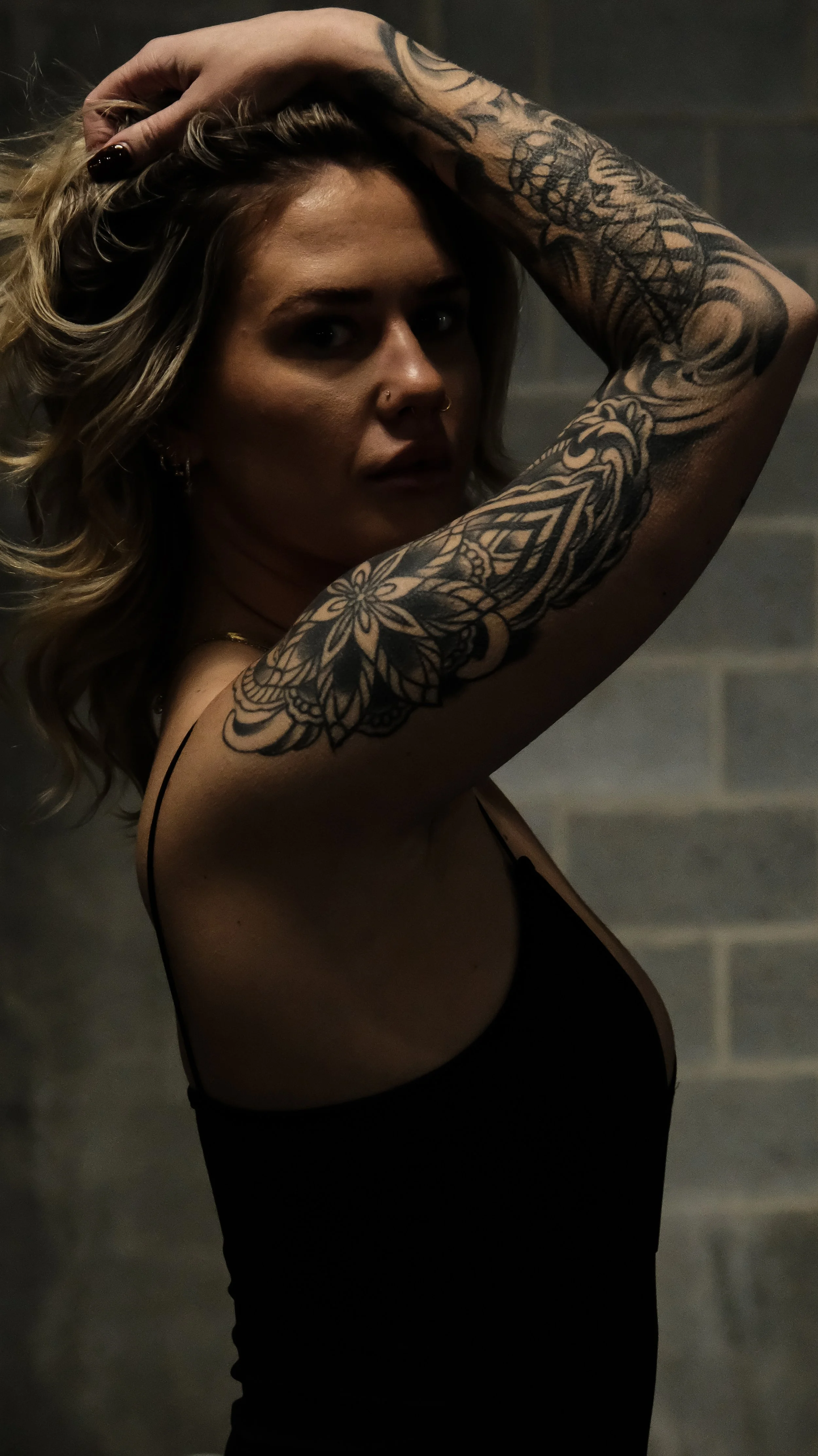 A woman with tattoos on her right arm, wavy blonde hair, and a nose piercing, posing with her hand on her head in front of a dark brick wall.