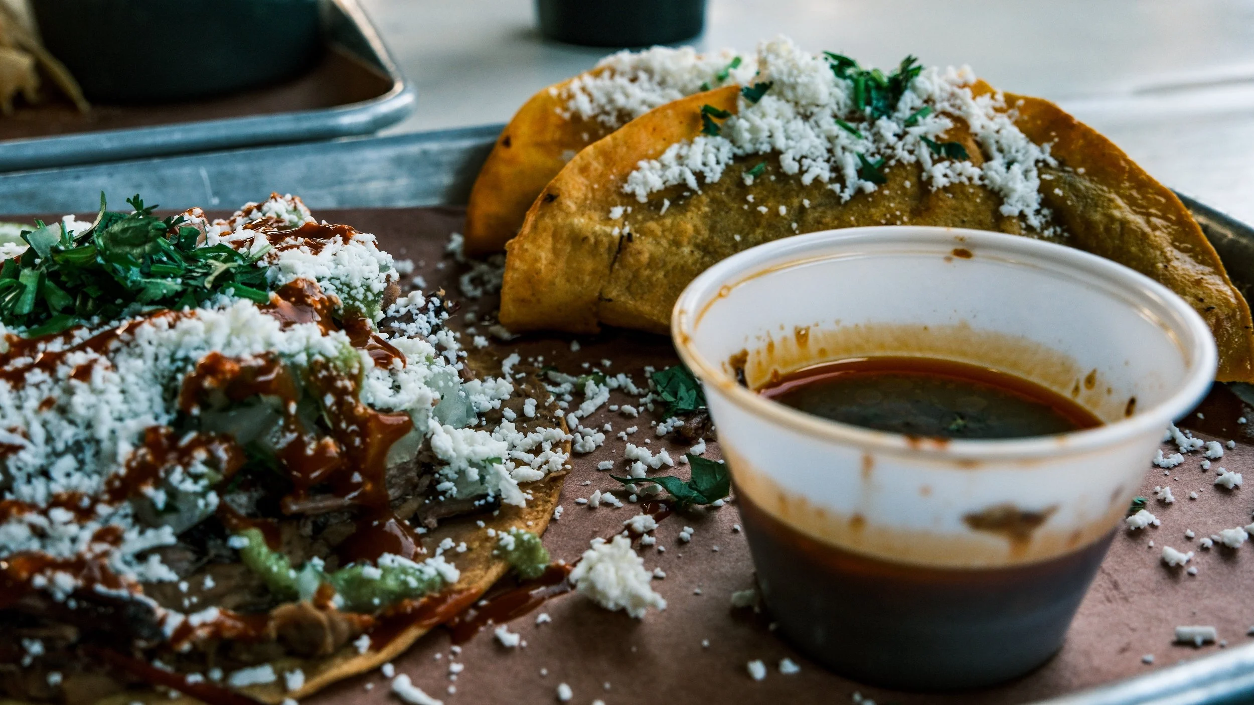 Three Mexican street tacos with shredded cheese, chopped herbs, and sauces on a tray, along with a cup of dark salsa.