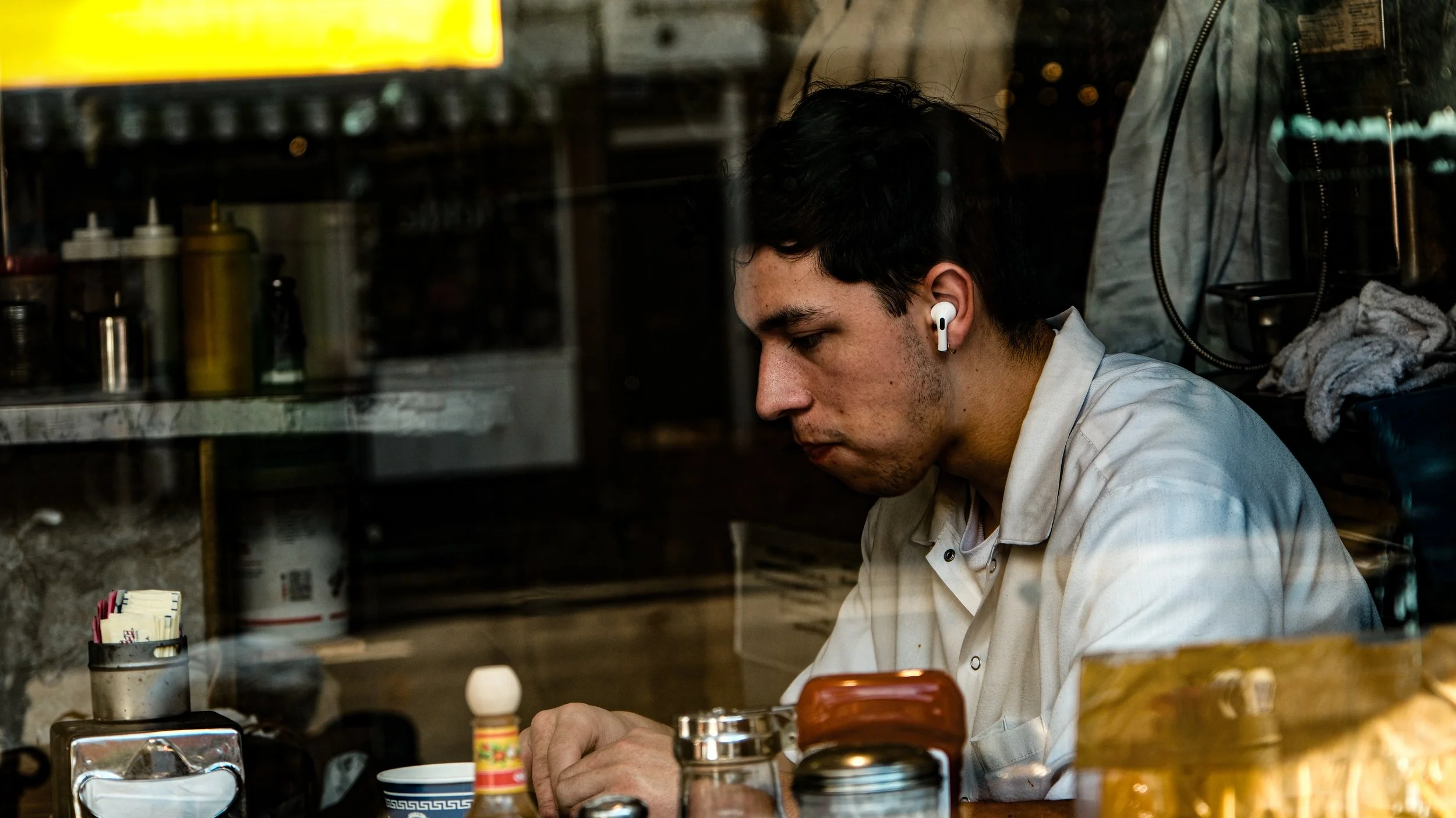 Young man sitting in a restaurant, wearing a white shirt and wireless earbuds, focused on his food or drink.