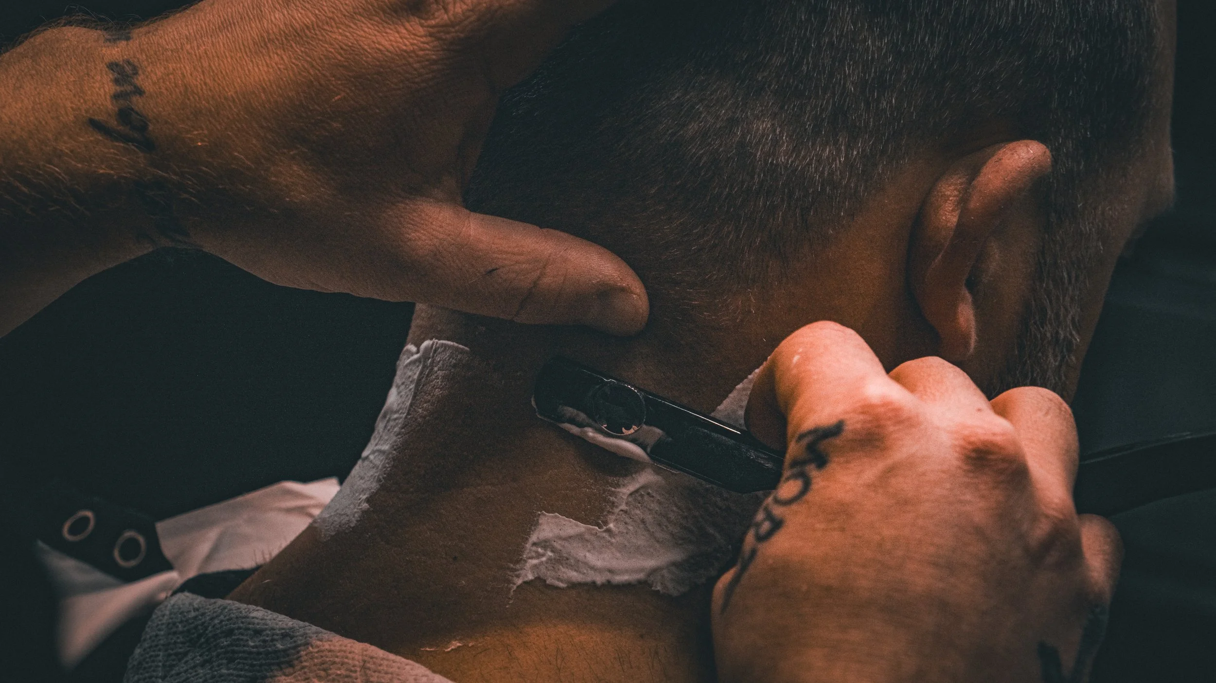A barber uses a straight razor to shave a man's neck, with white shaving cream on his skin.