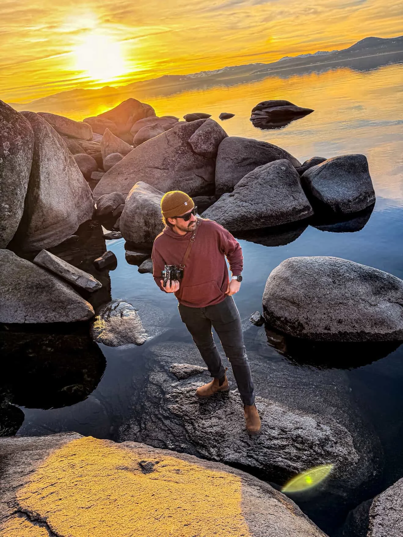 Man standing on rocks in a lake during sunset, holding a camera, wearing a red hoodie, sunglasses, brown boots, and a brown beanie.