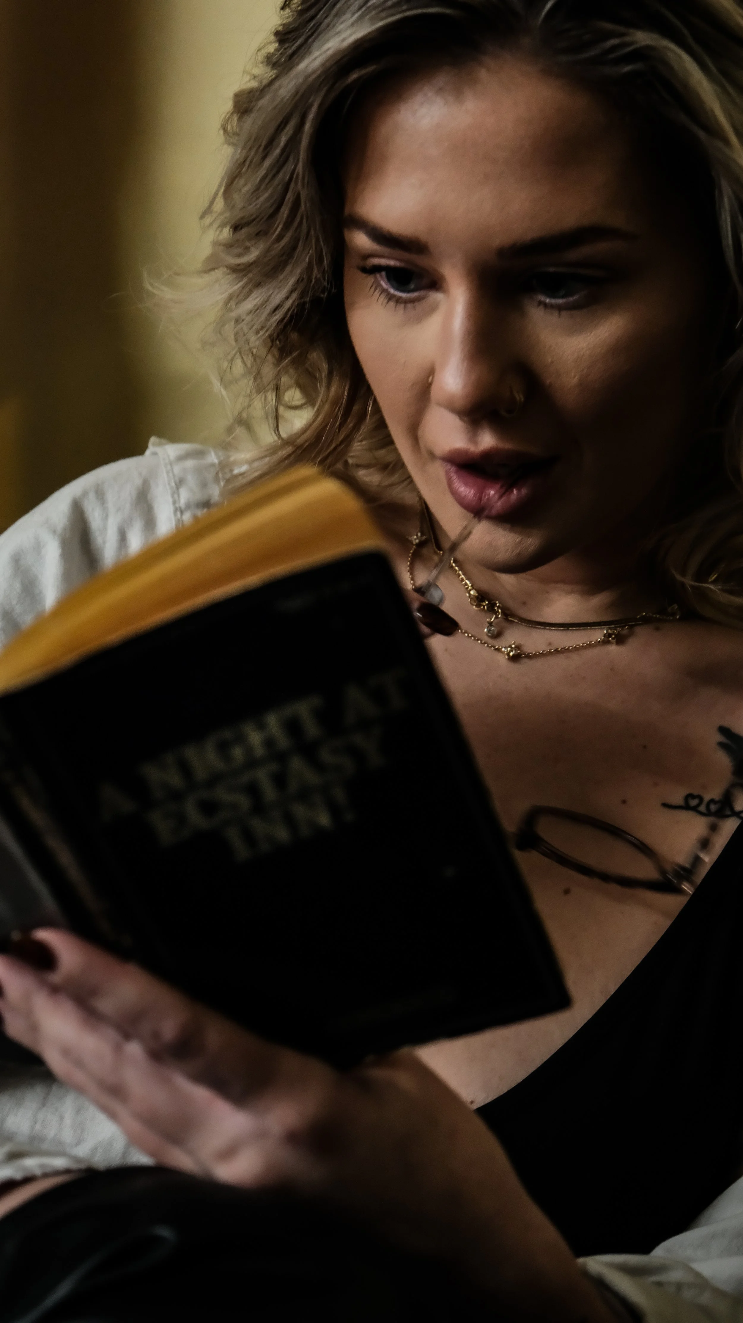 A young woman with wavy blonde hair, wearing layered necklaces and a white shirt, is looking at a book titled 'A Night at the East.
