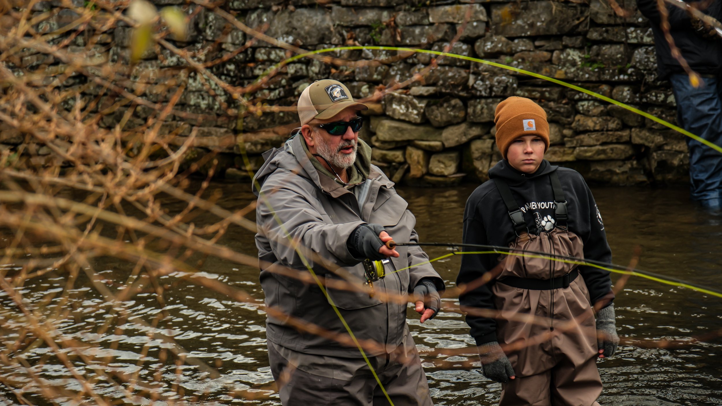 Two individuals fishing in a river, with one adult man wearing sunglasses and a baseball cap, and a young boy wearing a beanie, both dressed in waterproof gear.