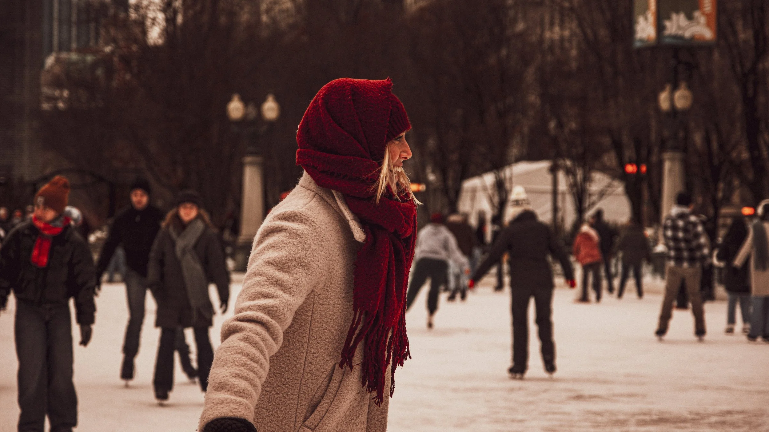 A woman wearing a red winter hat and scarf, with a beige coat, ice skating on an outdoor rink surrounded by other skaters during winter.