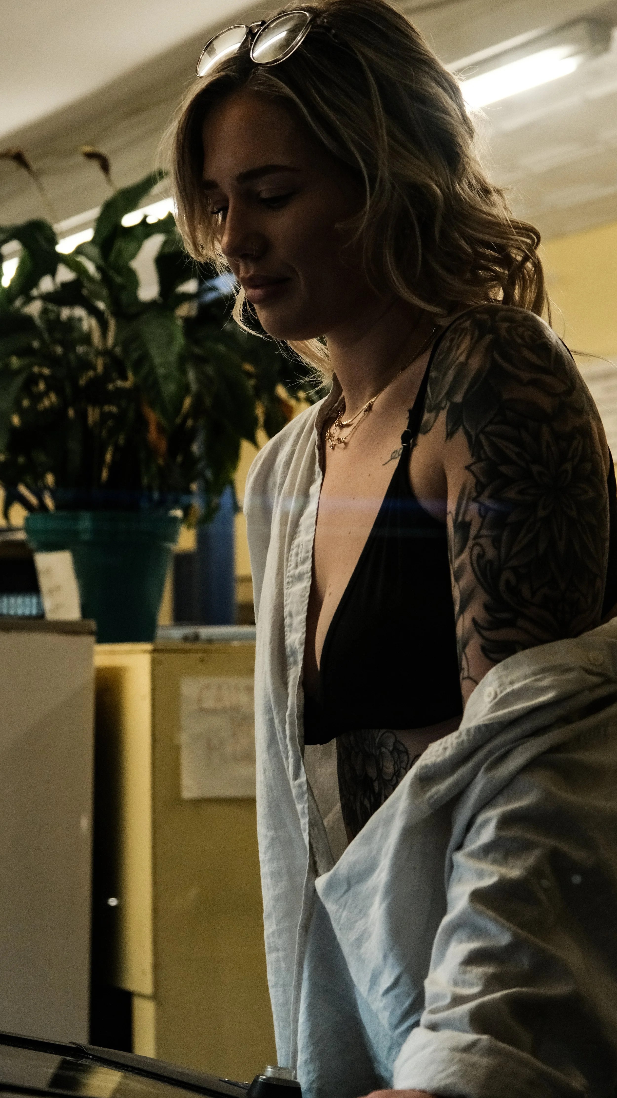 A young woman with wavy, shoulder-length hair and tattoos on her left arm, wearing glasses on her head, a black bra, and an unbuttoned white shirt, standing indoors with a large green potted plant in the background.