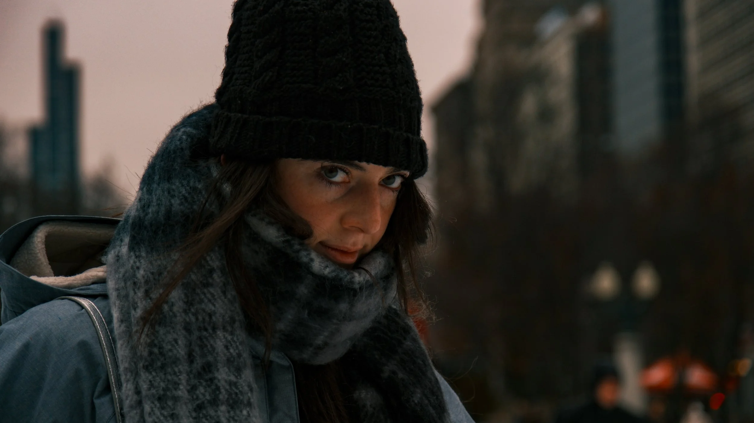 A woman with blue eyes wearing a black knitted beanie and a large grey scarf outdoors in an urban setting during dusk or dawn.
