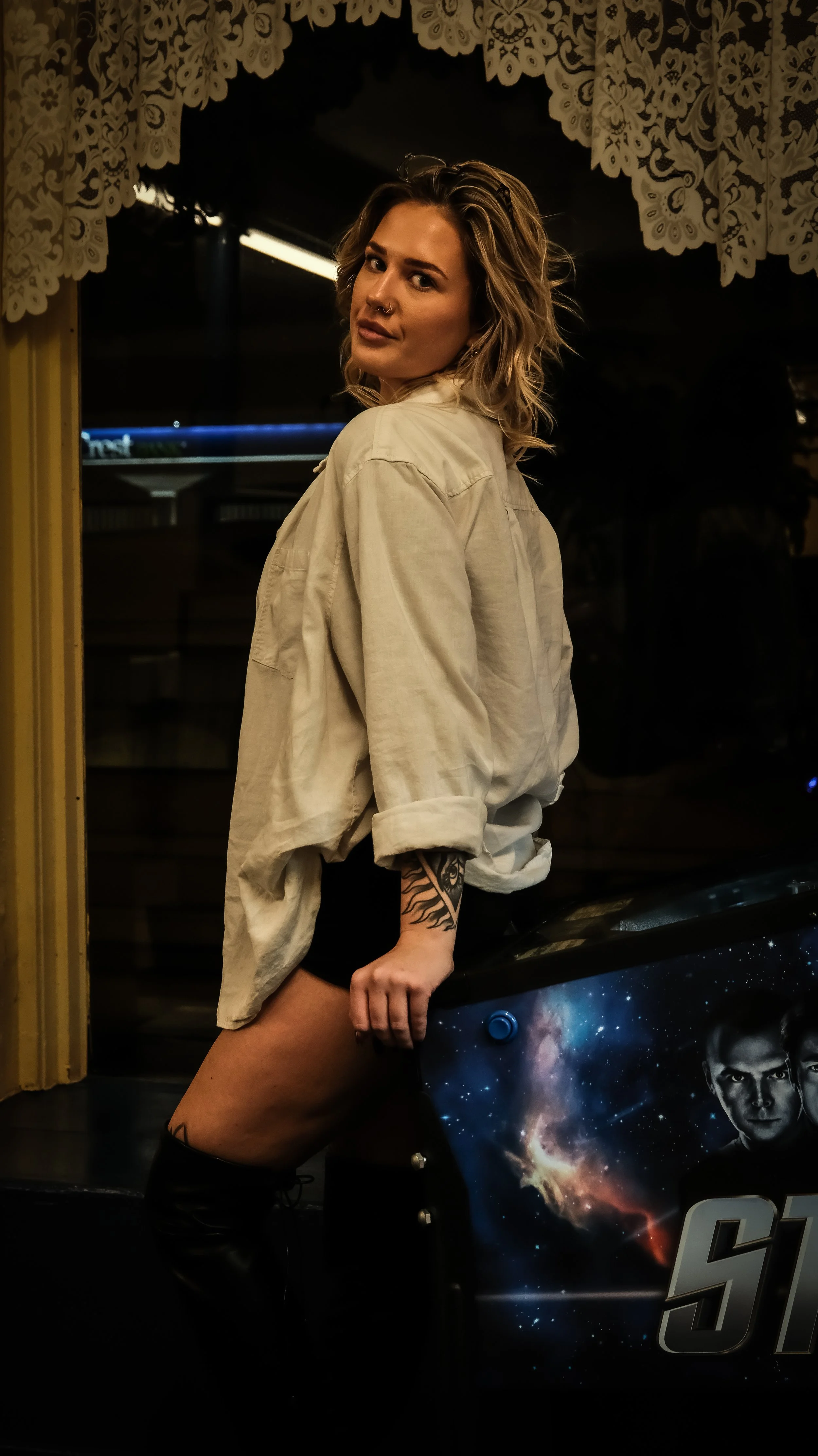 A woman with wavy blonde hair and tattoos on her arm and thigh leaning on a space-themed arcade game, wearing a beige oversized jacket and black shorts, in a dimly lit room.