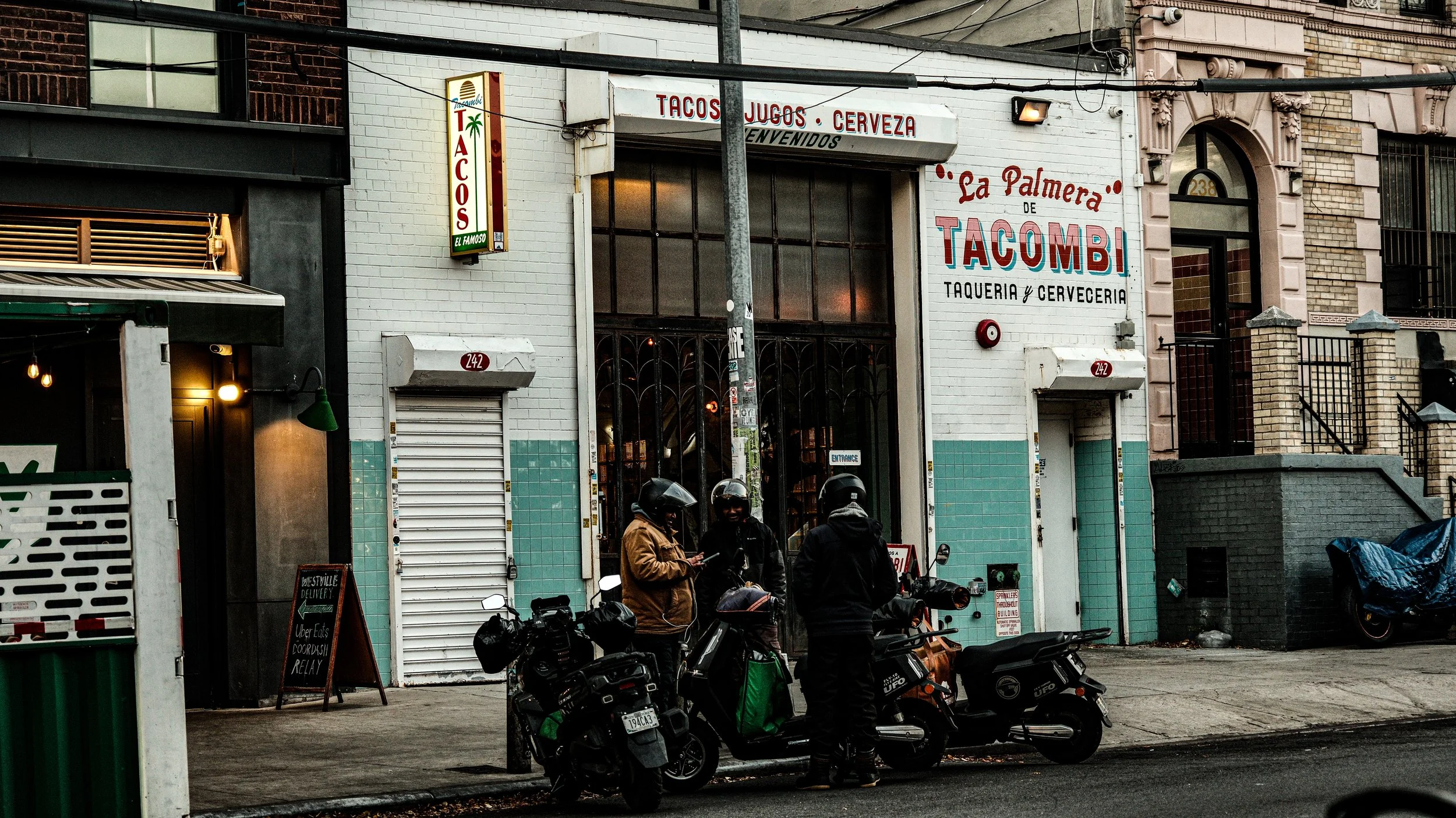 Three motorcyclists with helmets, one wearing a brown jacket, standing outside a Mexican restaurant called La Palmera de Tacombi, on a city street in front of a building with a white brick facade and large window.