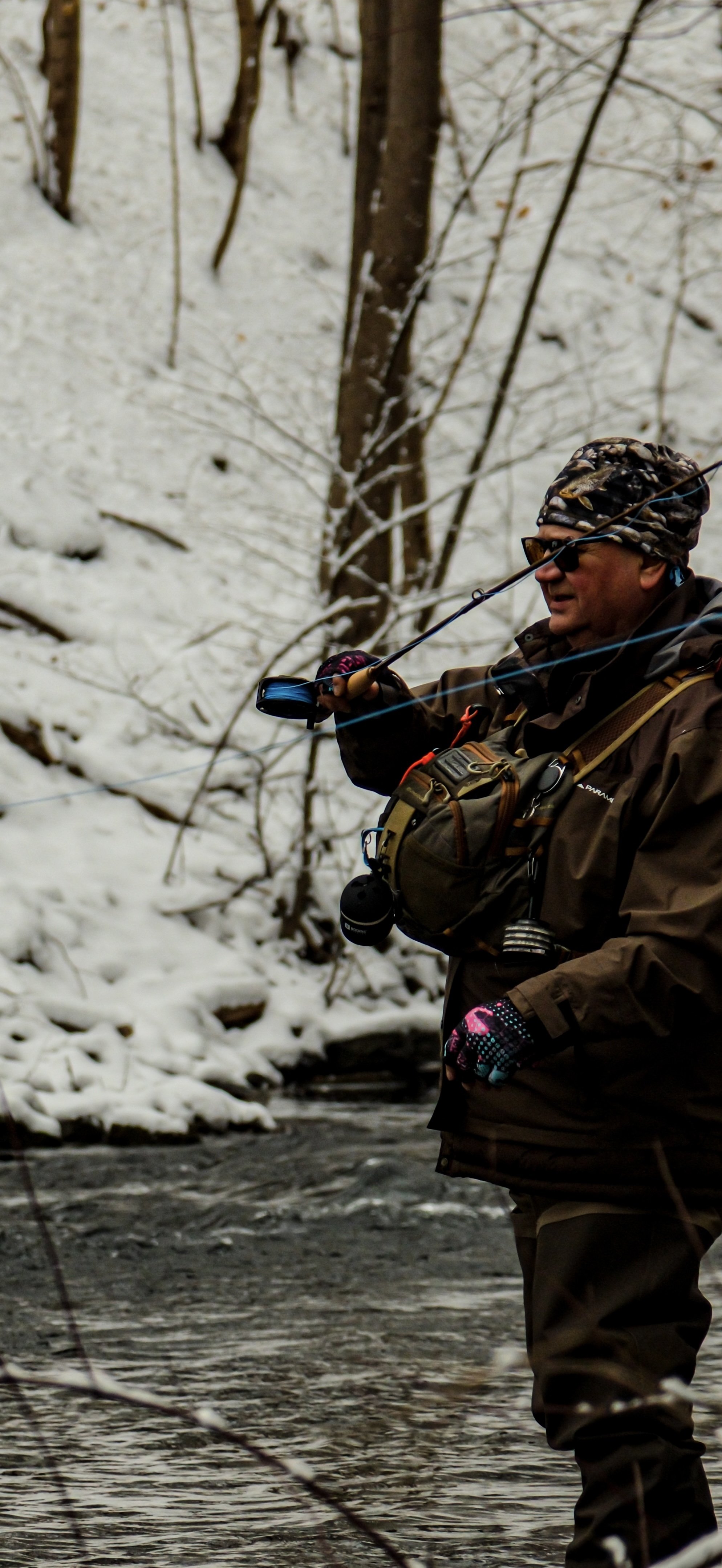 A man wearing sunglasses, a camouflage hat, and outdoor gear, standing in a snowy river while fishing with a spinning rod.