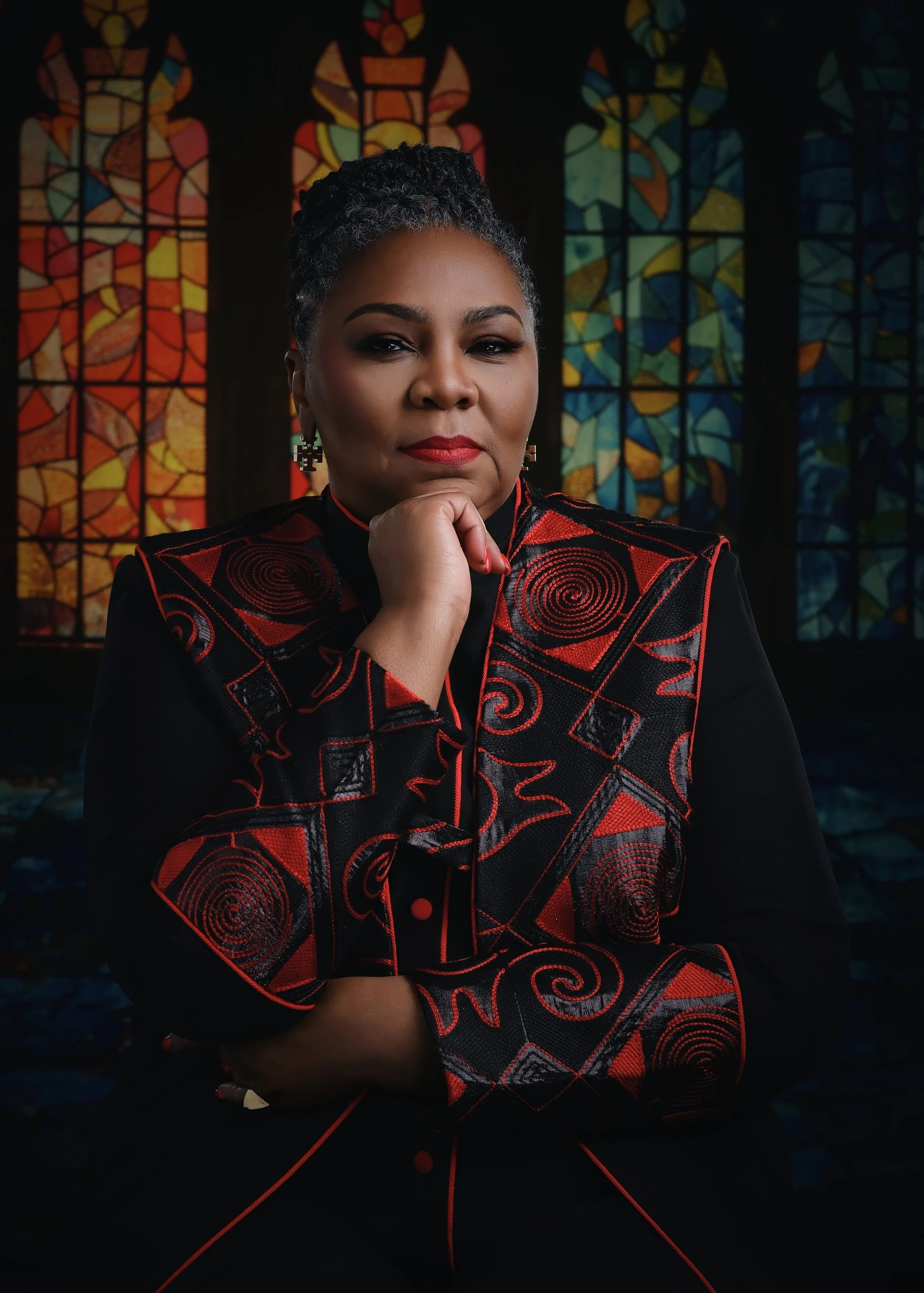 A woman with short grey curly hair, wearing a black and red embroidered jacket, sitting in front of stained glass windows with colorful abstract patterns, resting her chin on her hand and looking confident yet welcoming.