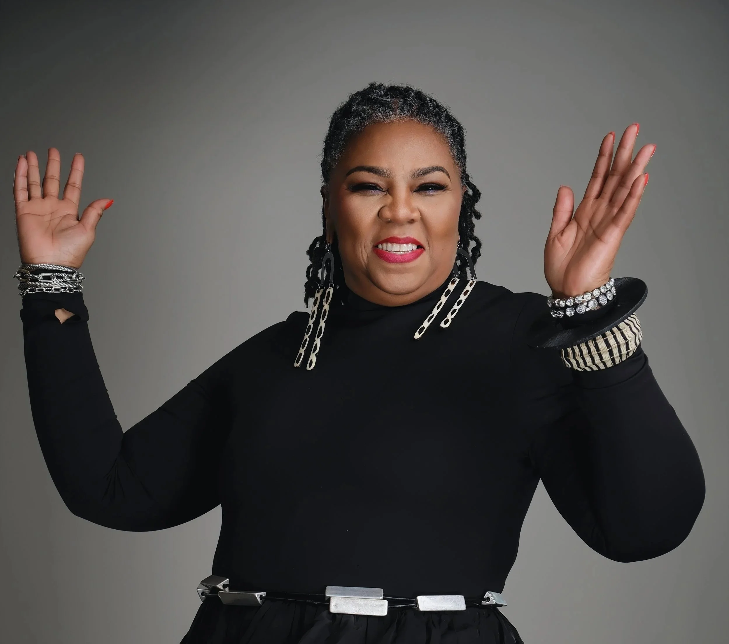 A woman with short curly hair wearing a black dress with layered silver jewelry and with her hands raised up, making a joyful expression.