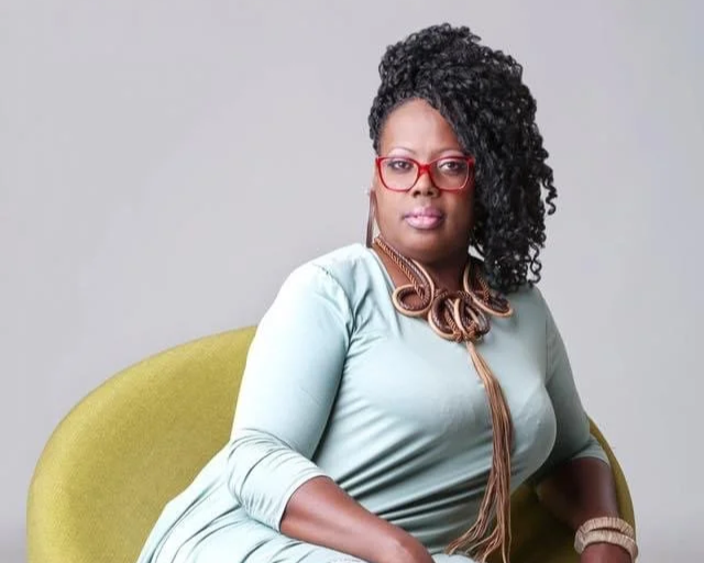 A woman with curly hair, red glasses, and red lipstick sitting on a yellow chair, wearing a light blue top and a large statement necklace with brown accents.