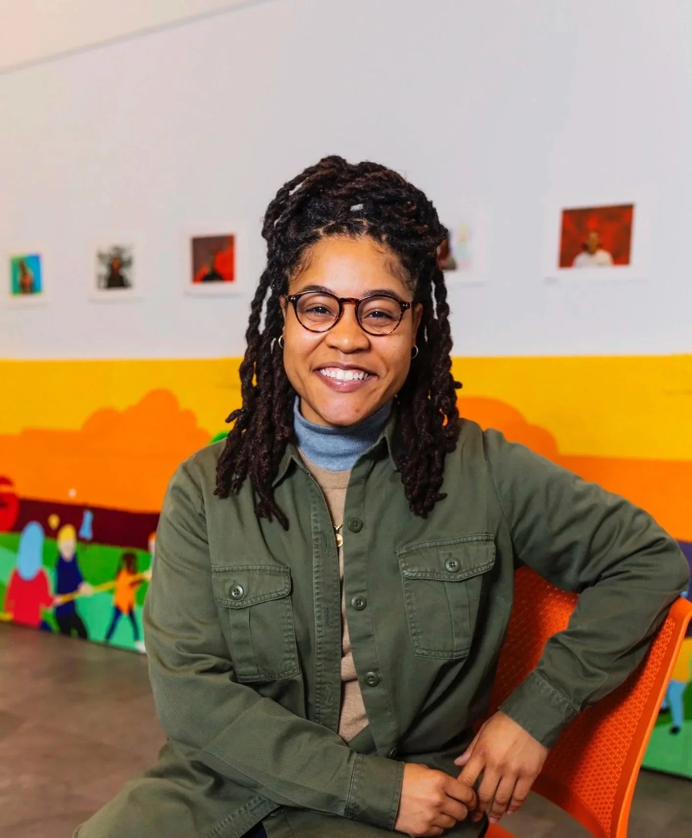 A woman with glasses and dreadlocks smiling at the camera, sitting on an orange chair, with colorful wall art behind her.