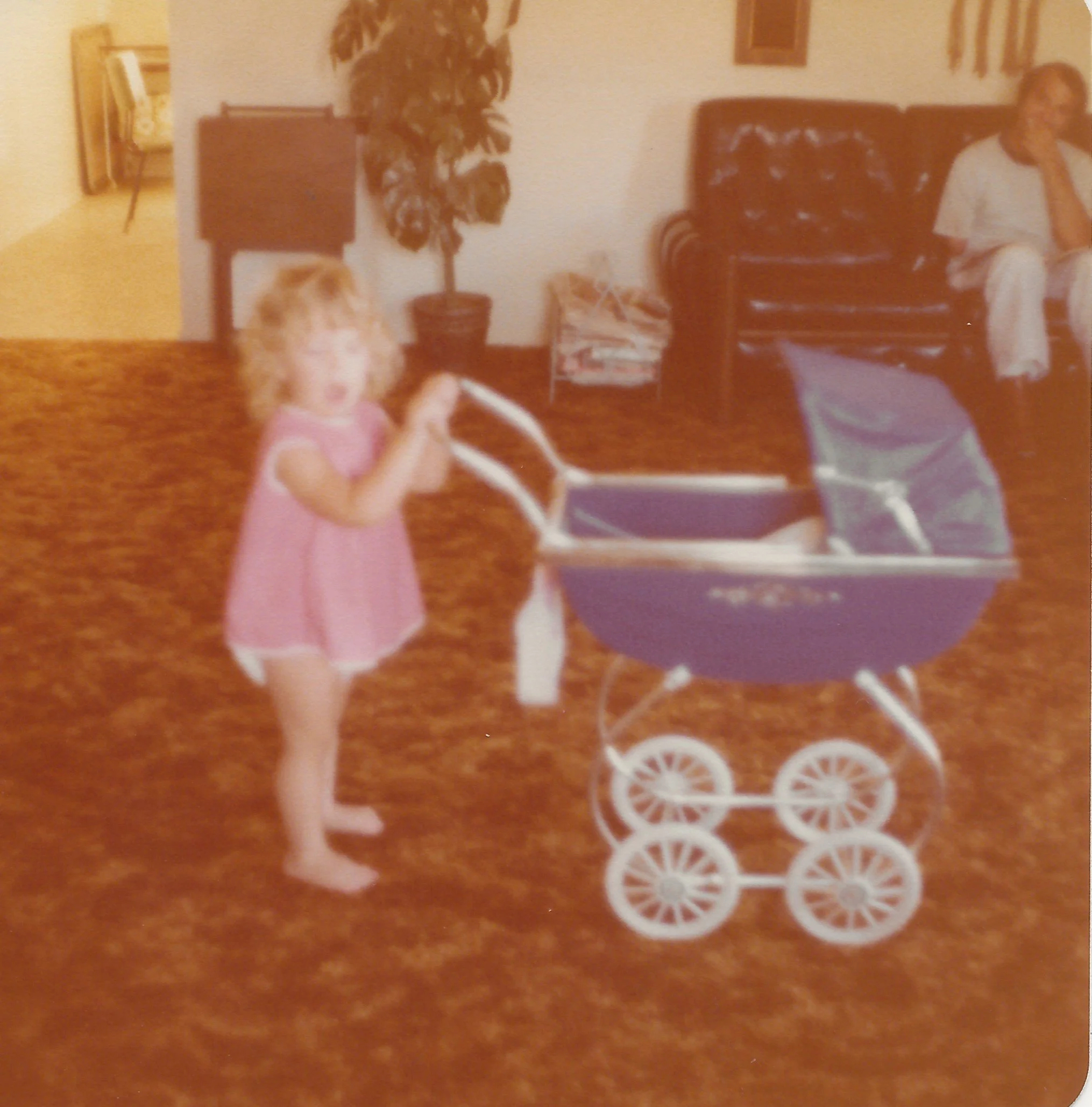 A woman and a young girl standing in a living room, with a dog sitting in front of the girl. The woman is seated on a dark brown leather couch, and there is a potted plant and framed pictures on the wall behind them. A box and some objects are on a small table in the background.