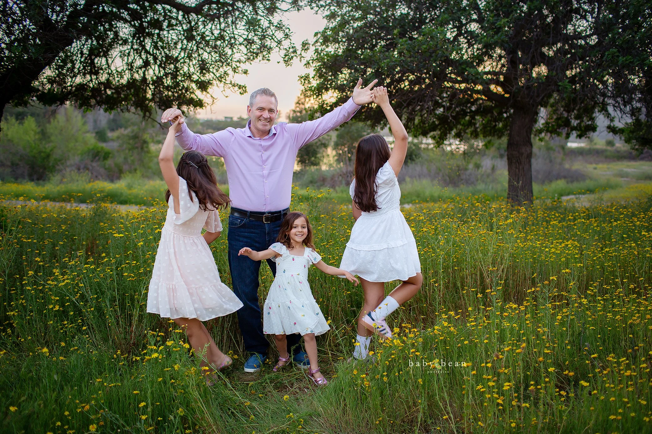 A smiling man in a purple shirt and four young girls in white dresses dancing and playing in a field of yellow flowers under large trees during sunset.