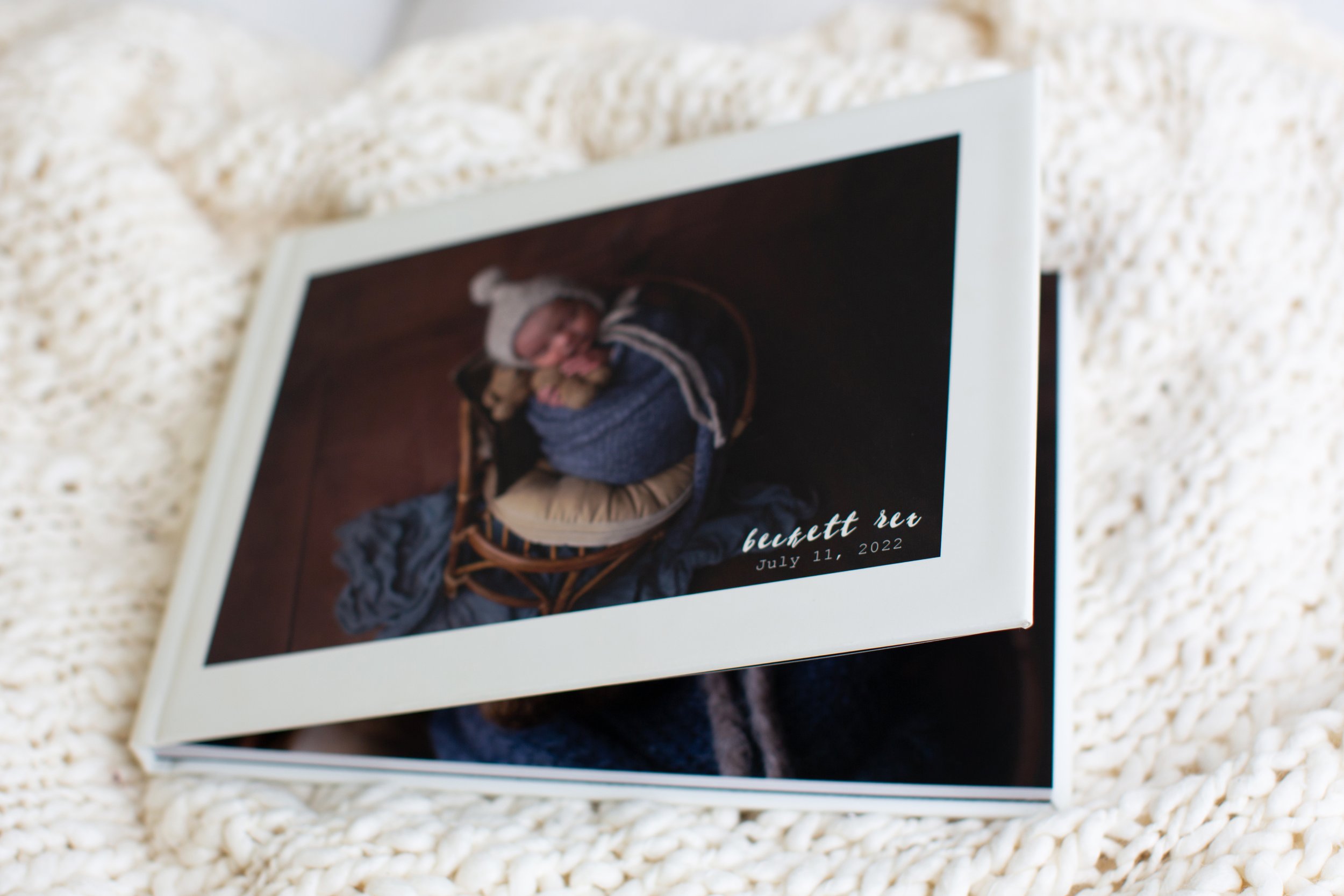 Close-up of a printed photo capturing a cozy scene of a child sleeping in a basket, wrapped in blankets, with the text overlay indicating a bucket set date of July 11, 2022, placed on a textured, cream-colored surface.