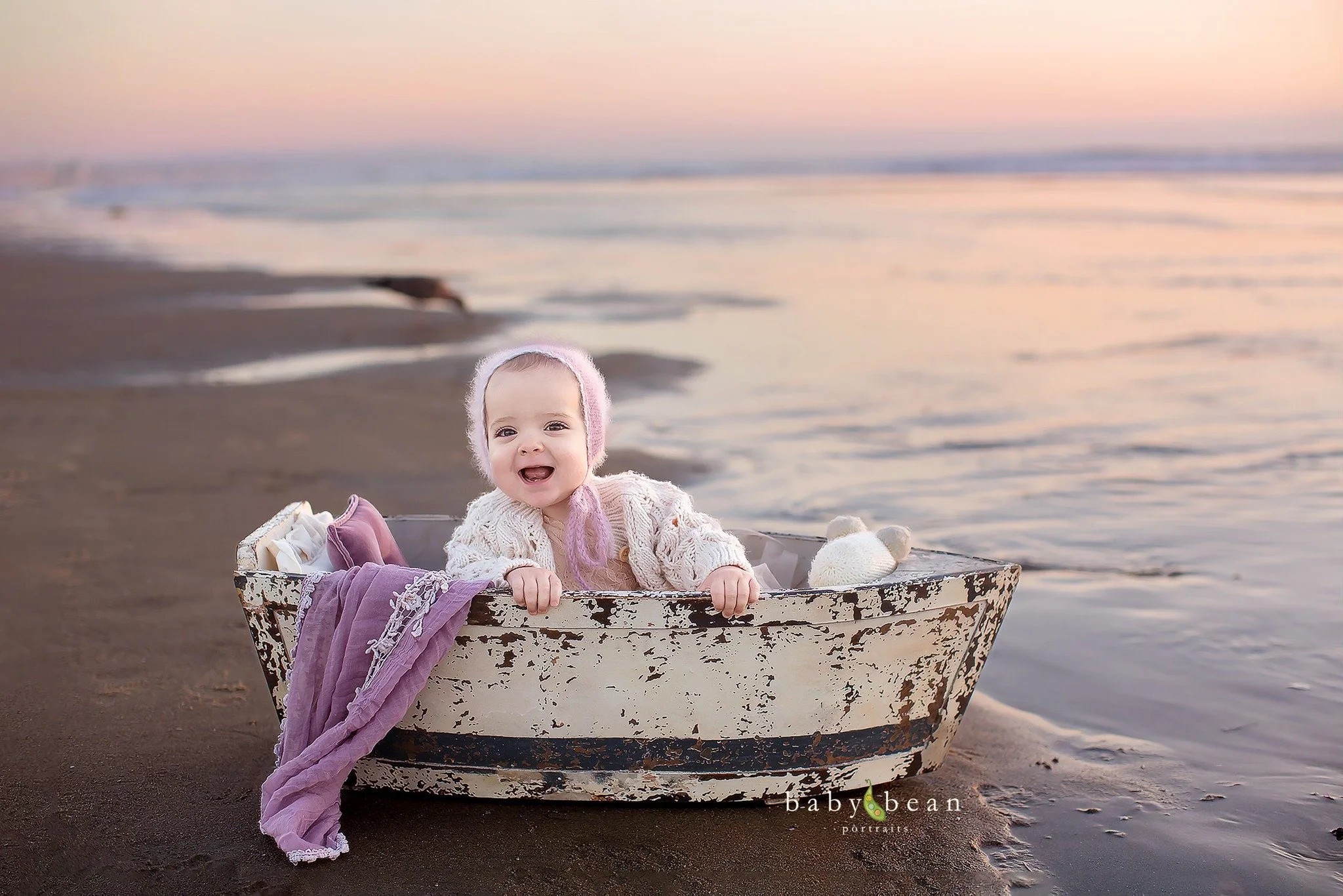 A smiling baby in a knitted cardigan and pink hat sitting in an old rowboat on the beach at sunset, with a purple blanket draped over the side and soft waves in the background.