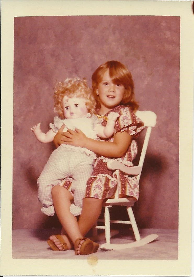 A young girl with red hair wearing a patterned dress, sitting on a white rocking chair, holding a curly-haired doll.