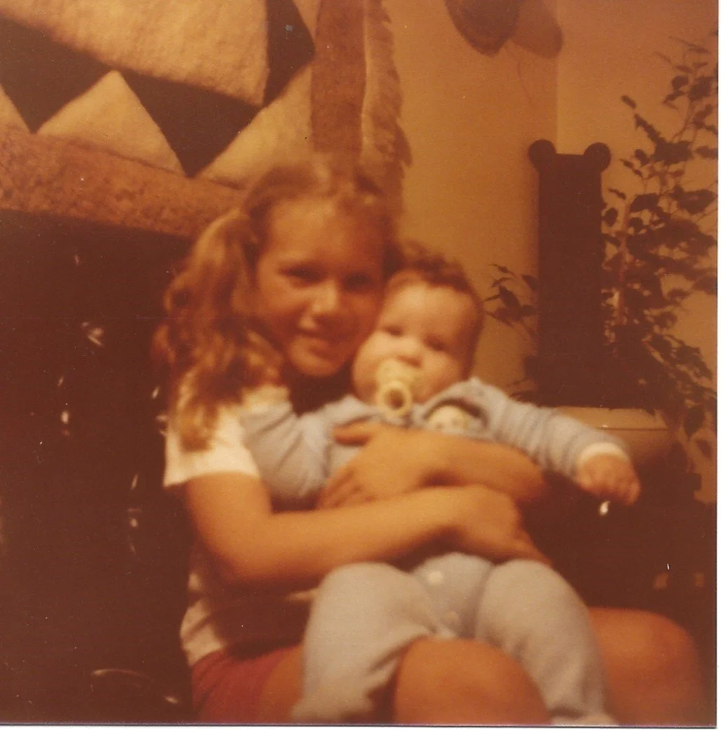 A young girl with long hair holding a baby with a pacifier in its mouth, sitting on a black chair in a cozy living room.