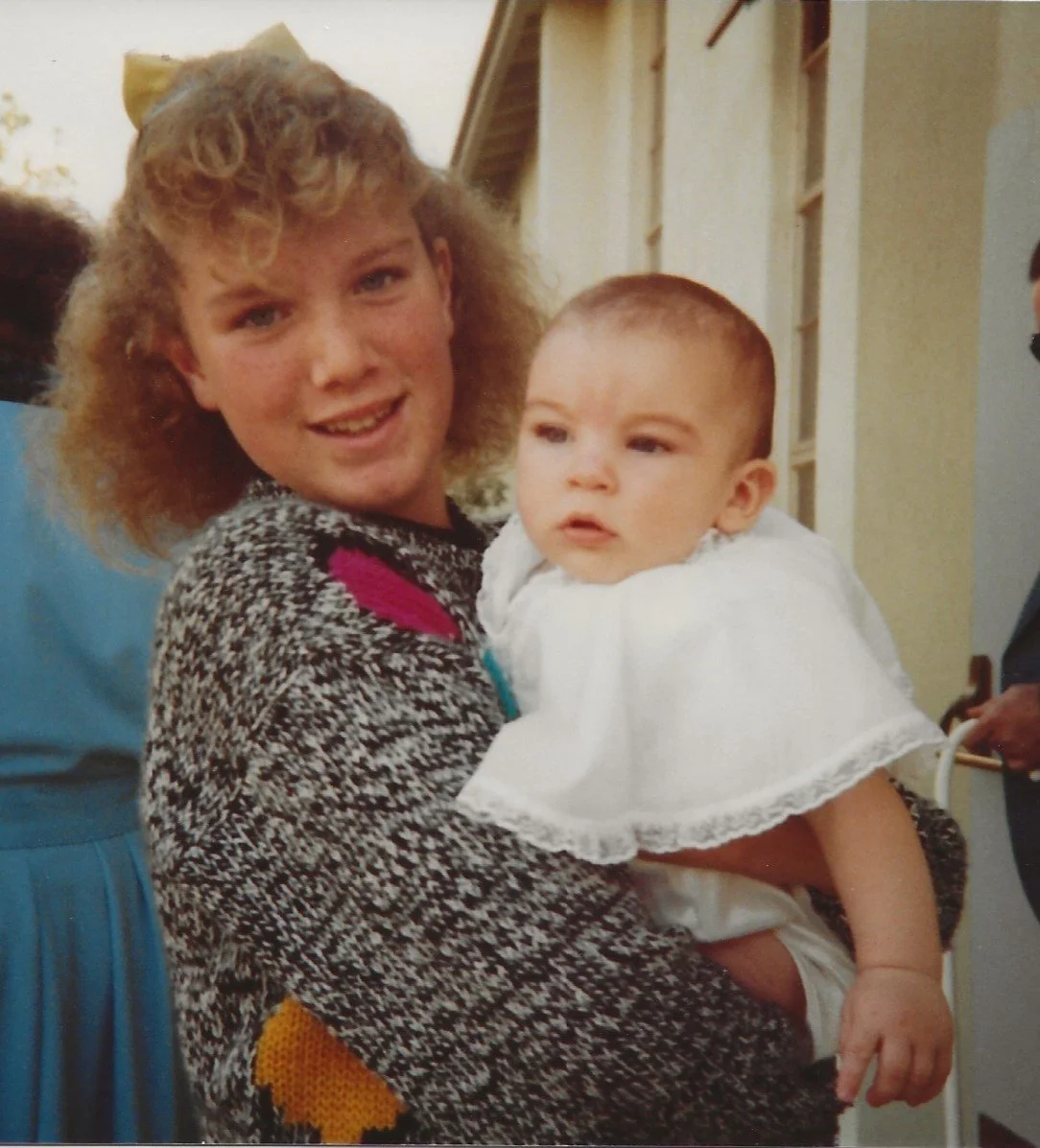 A woman with curly blonde hair holding a young child dressed in a white garment, both smiling at the camera outside a house.