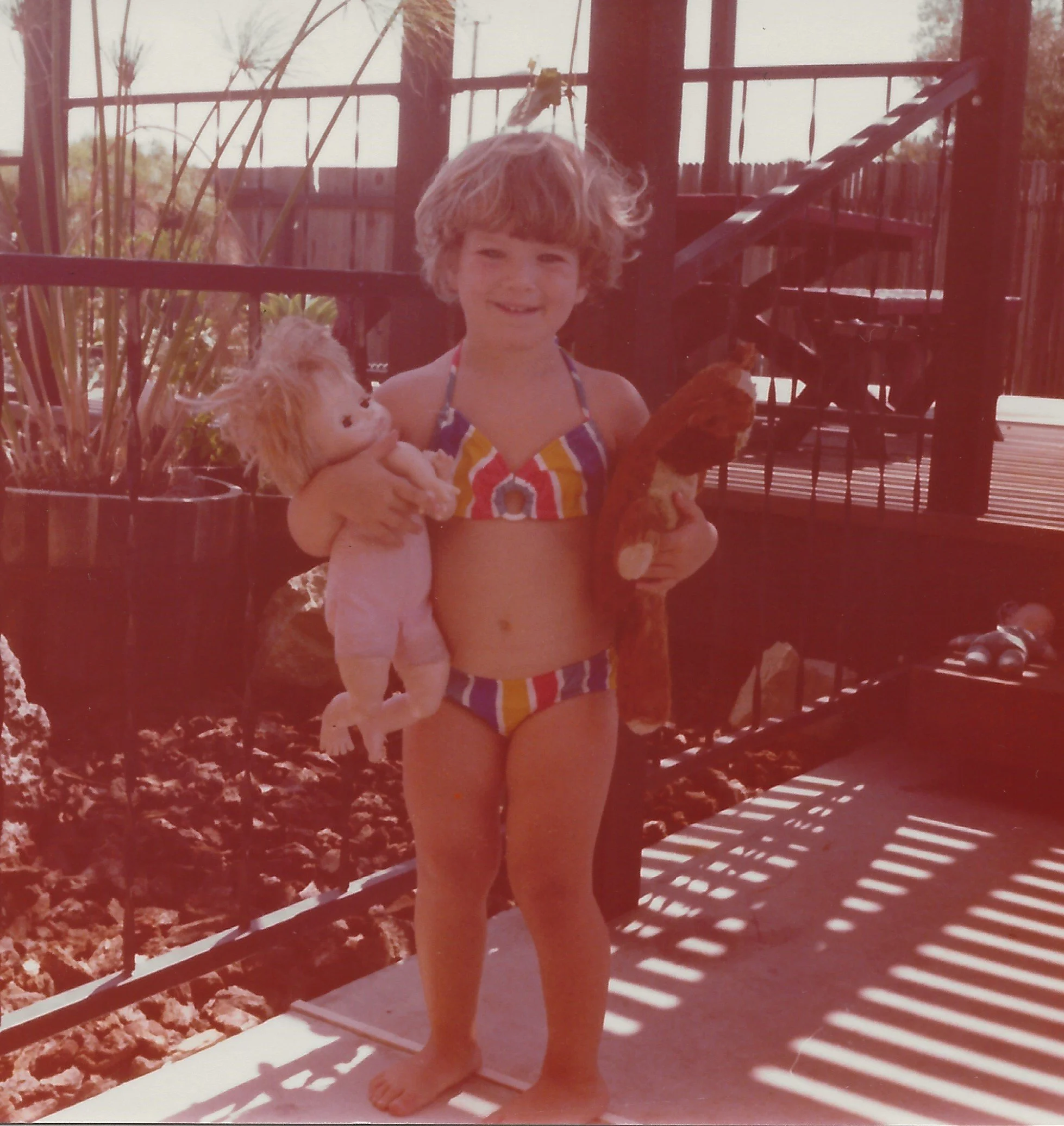 Child in colorful swimsuit holding plush lion toy, standing on balcony with wooden floor, shadows cast by railing, outdoor trees in background.