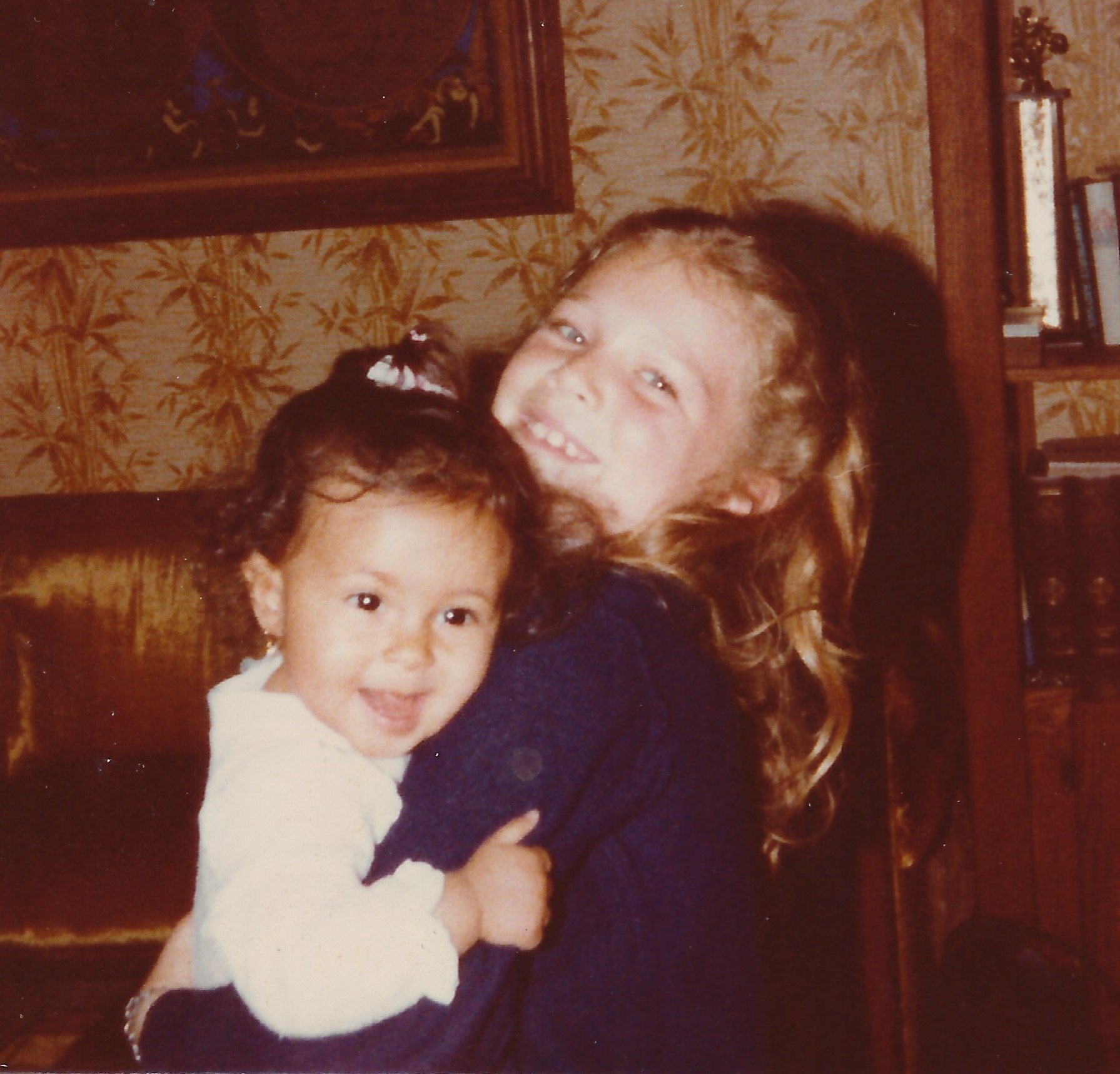 A young girl with curly hair and light skin hugging a smaller child with straight dark hair and light skin in a room with wooden furniture and framed pictures on the wall.