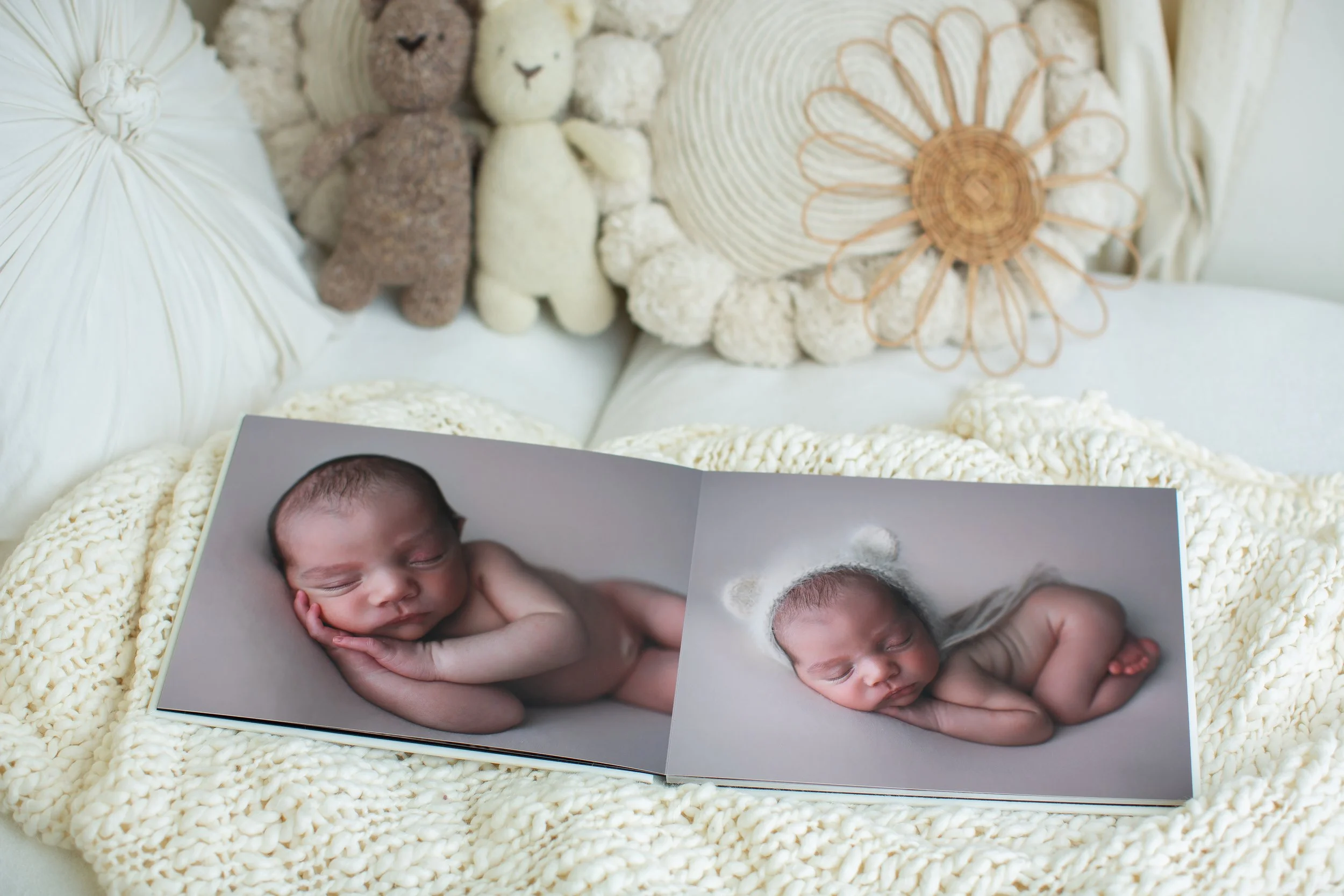Close-up of a photo album with images of a sleeping baby, placed on a fluffy white knitted blanket, surrounded by plush toys, decorative pillows, and a woven sun-shaped wall decor.