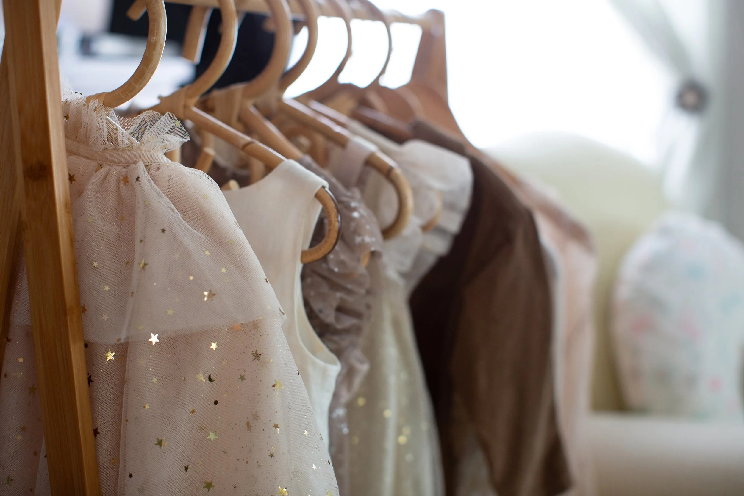 A row of children's dresses hanging on wooden hangers on a wooden rack, with some dresses featuring star patterns and tulle fabric, in soft pastel and neutral tones.