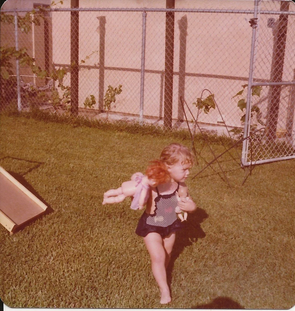 A young girl with curly red hair running on a lawn, holding a doll in her hand, with a fenced yard and a slide in the background.