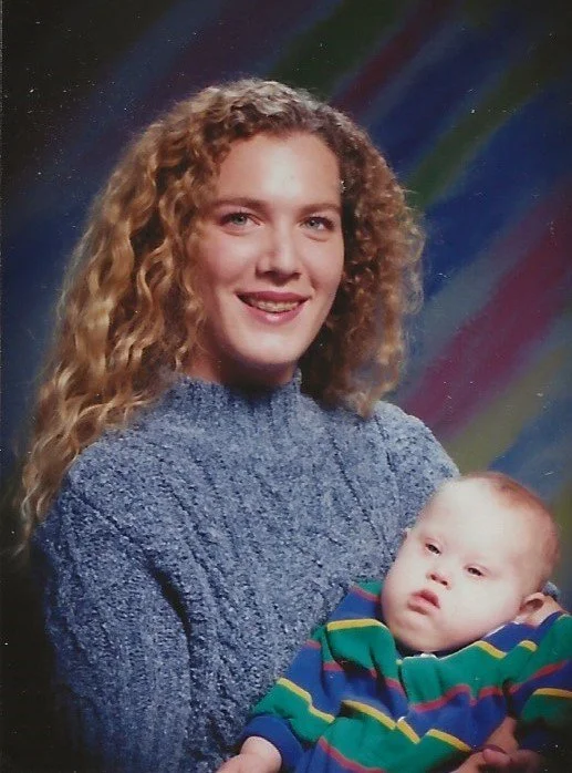 Young woman with curly blonde hair smiling and holding a young child with light hair wearing a colorful striped shirt, against a rainbow-colored background.
