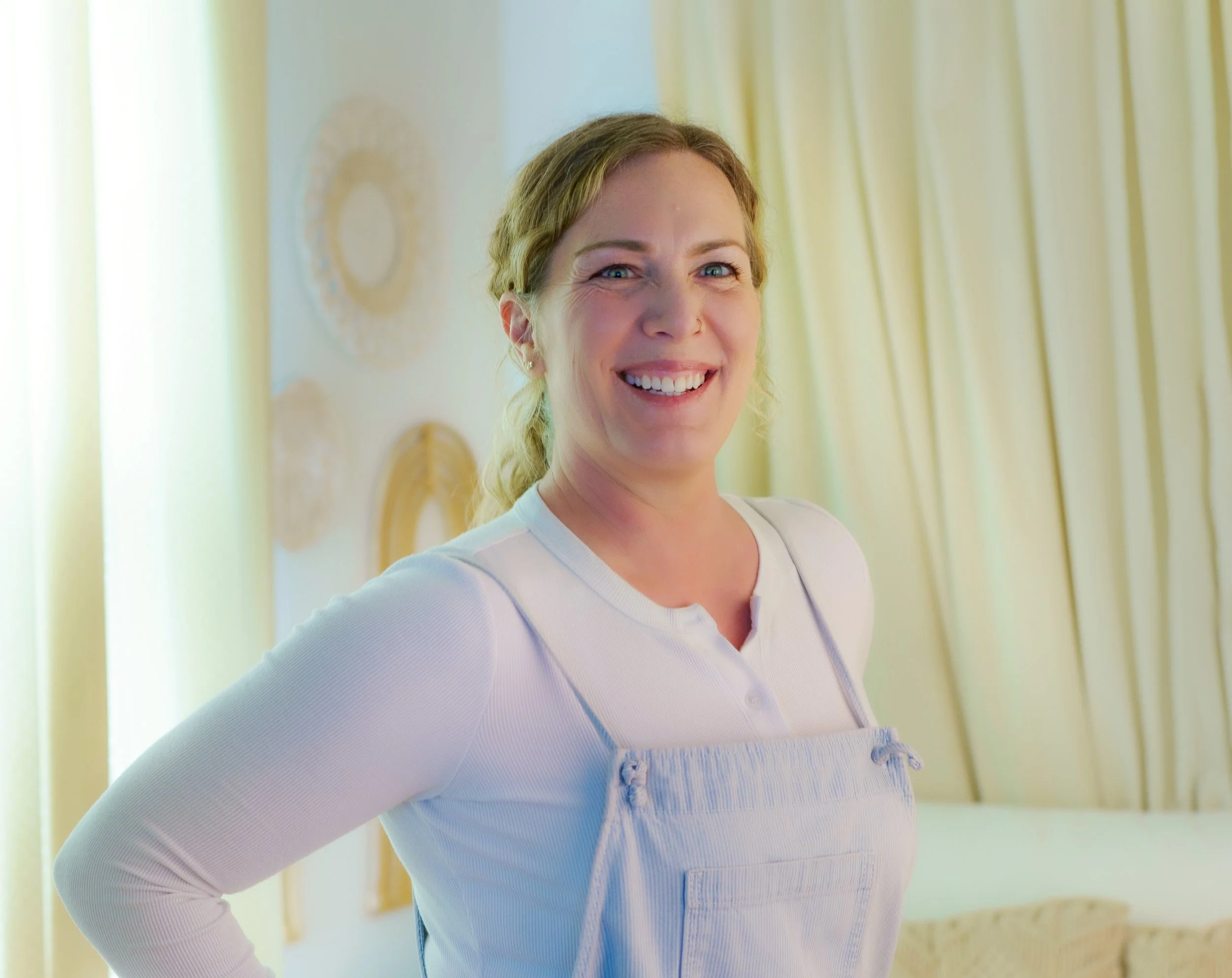 Smiling woman with blonde hair in a white top standing indoors with light-colored curtains behind her.
