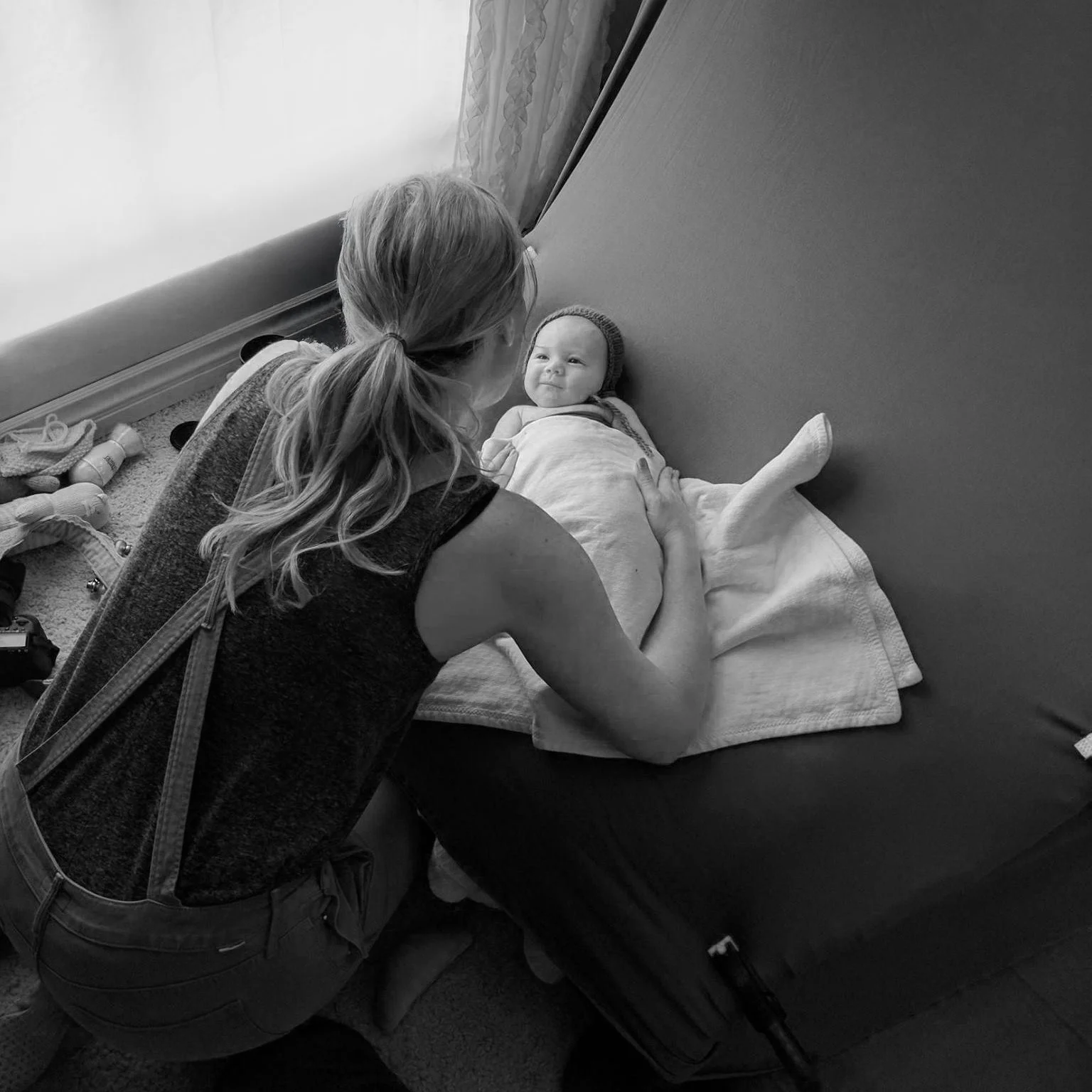A woman with long hair in a ponytail leaning over a baby who is lying on a bed. The baby is wearing a hat and wrapped in a blanket, looking at the woman. The room has a window with curtains.