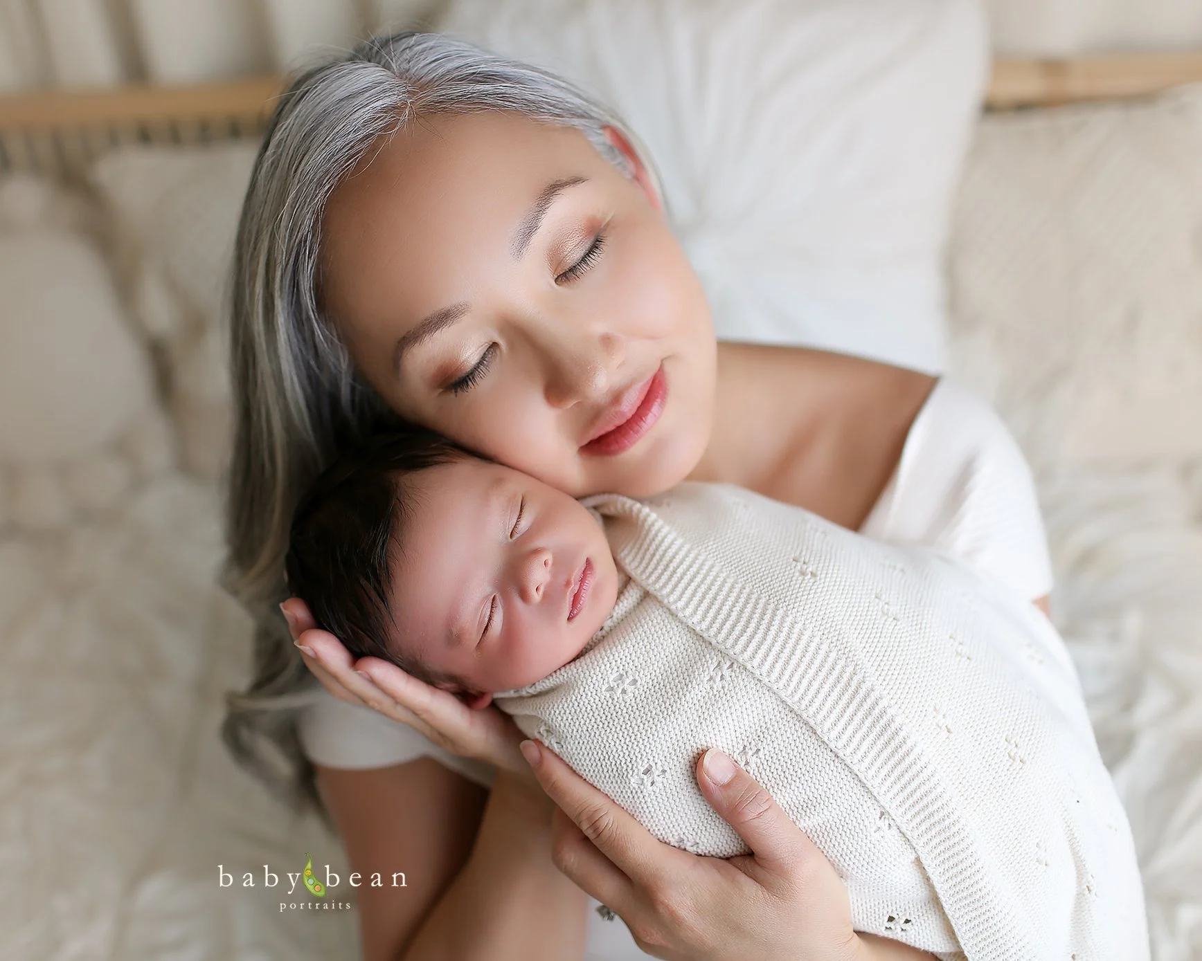 A woman with gray hair cradling a sleeping newborn baby wrapped in a cream-colored knitted blanket, both resting peacefully on a bed.