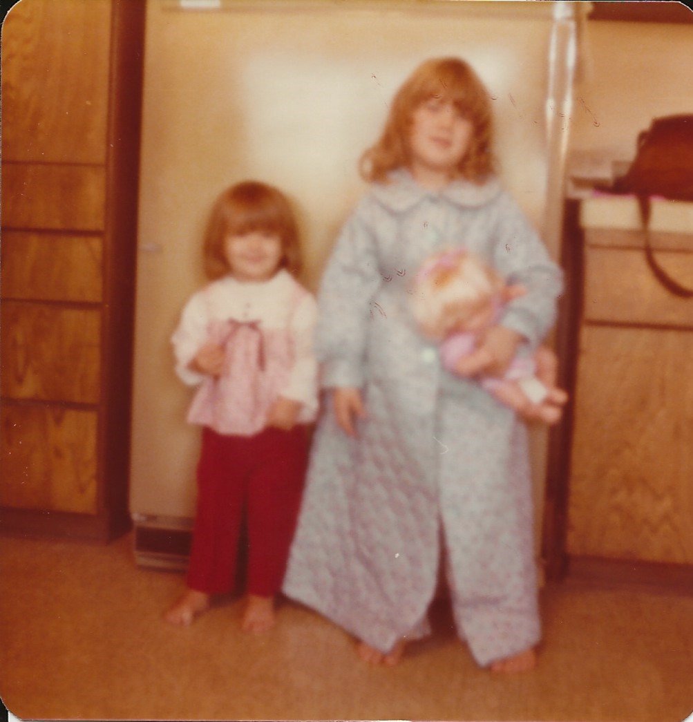 Two young girls with red hair standing on a carpeted floor indoors, holding dolls. One girl is shorter, wearing a white top with pink accents and red pants, while the taller girl is in pajamas with a floral pattern, holding a doll.