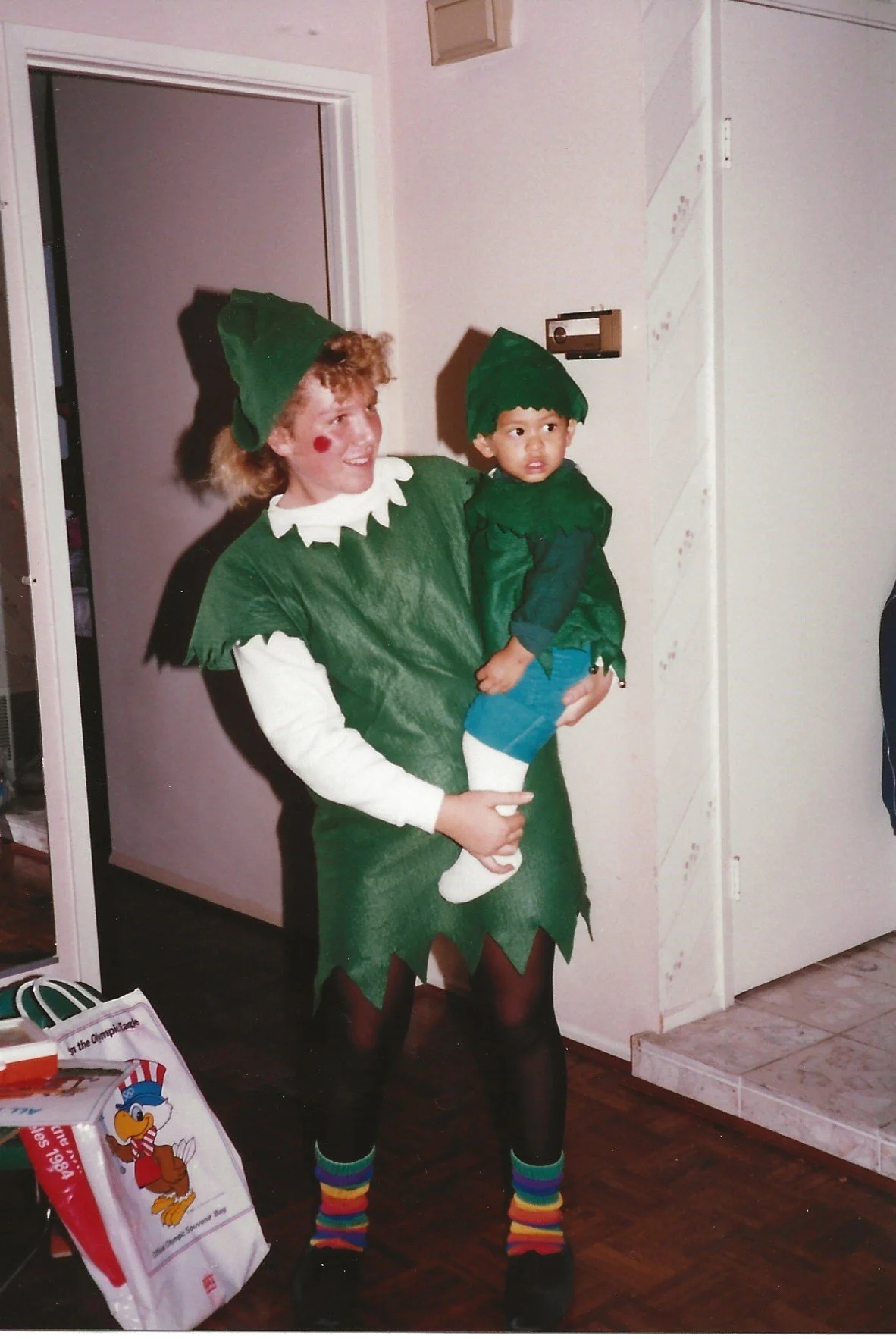 A woman dressed as a Christmas elf holding a young boy also dressed as an elf in a home interior setting. The woman is wearing a green elf costume with a matching hat, and the boy is wearing a green elf outfit with a hat. Both are wearing colorful striped socks.