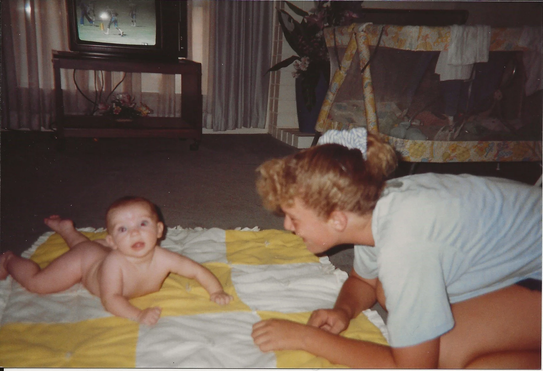 A baby lying on a bed with a yellow and white blanket, looking at the camera. An adult woman with short curly hair and a light-colored shirt is leaning close, smiling at the baby. In the background, a television displaying a sports game, a flowered plant in a vase, and a patterned sofa with towels or cloth on it are visible in a room with curtains and a window.