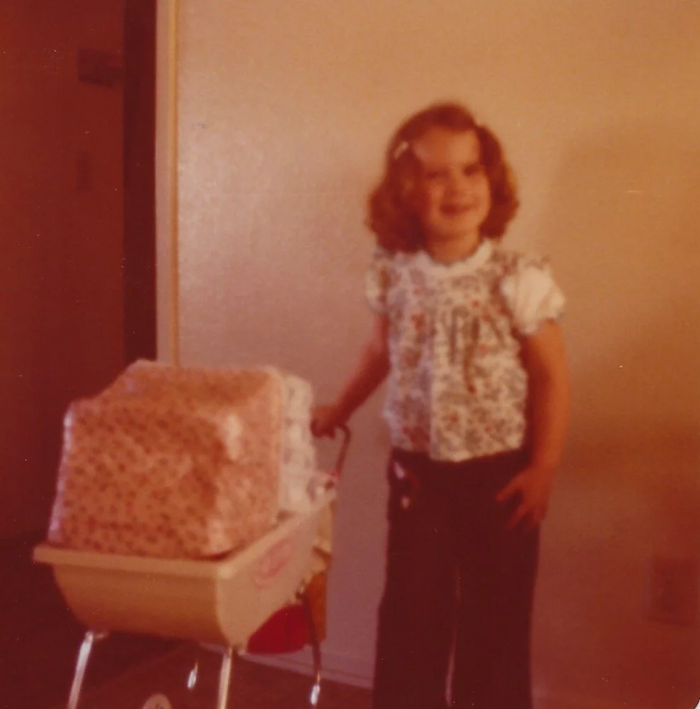 A young girl with curly hair, wearing a floral top and dark pants, smiling and standing next to a toy stroller with a large wrapped gift on top.