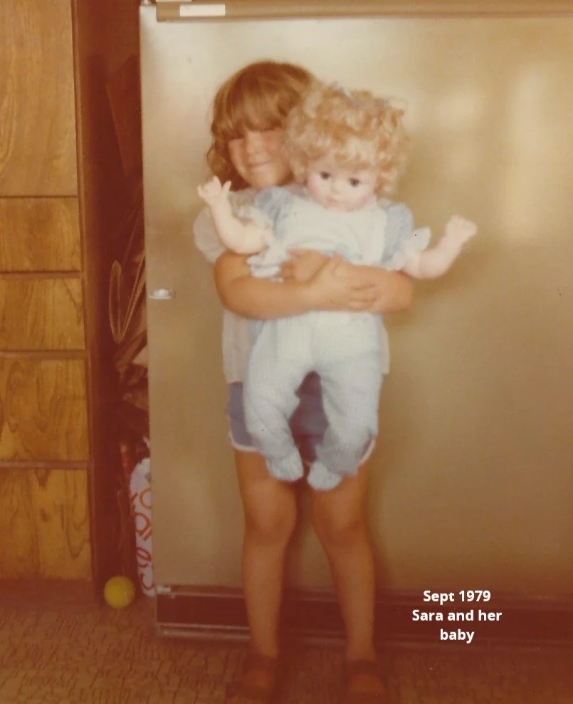 A young girl with curly hair holding a small curly-haired baby in front of a beige refrigerator in a kitchen. Text on the image reads, 'Sept 1979 Sara and her baby'.