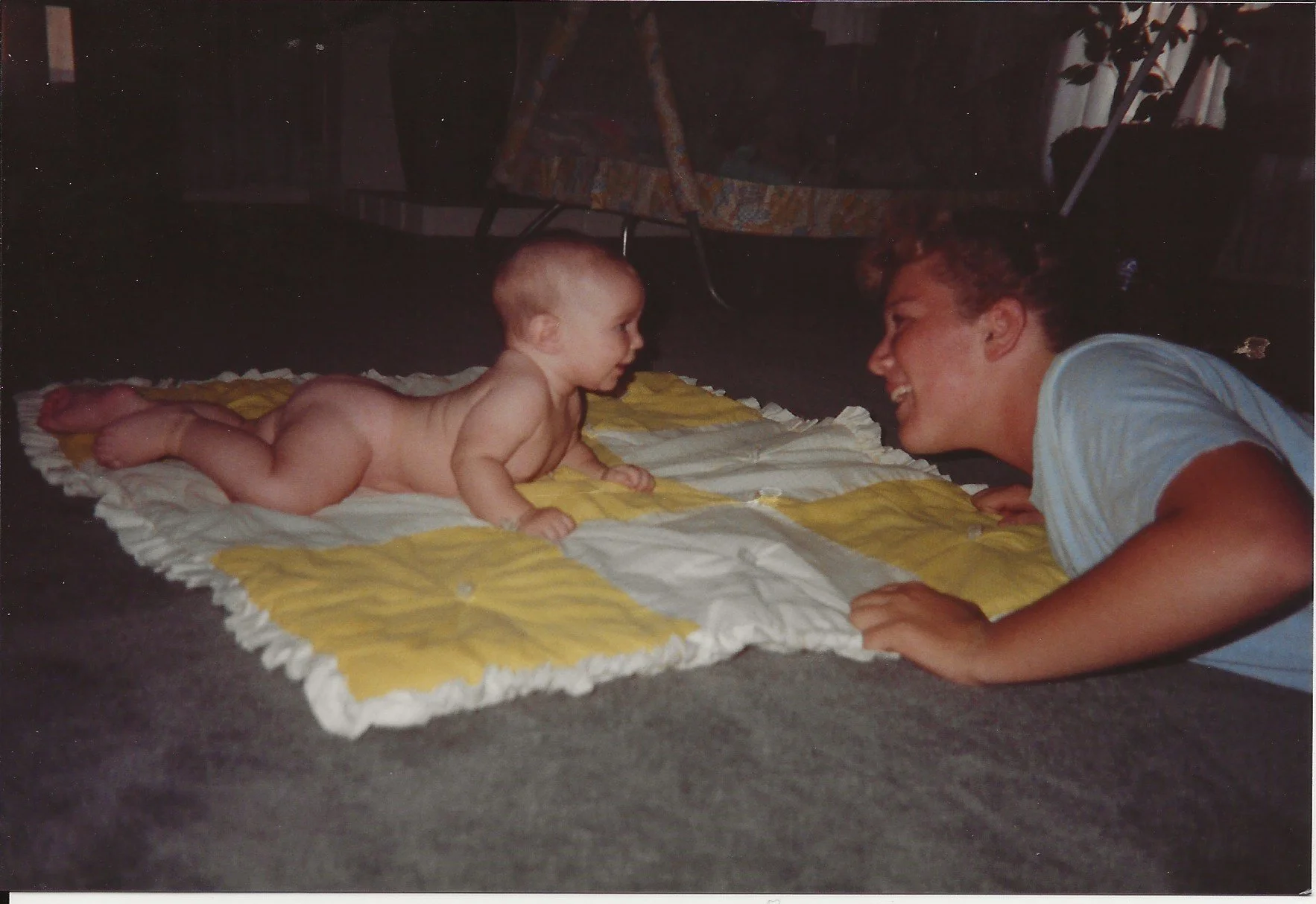 A woman and a baby lying face to face on a quilted blanket, smiling and looking at each other.
