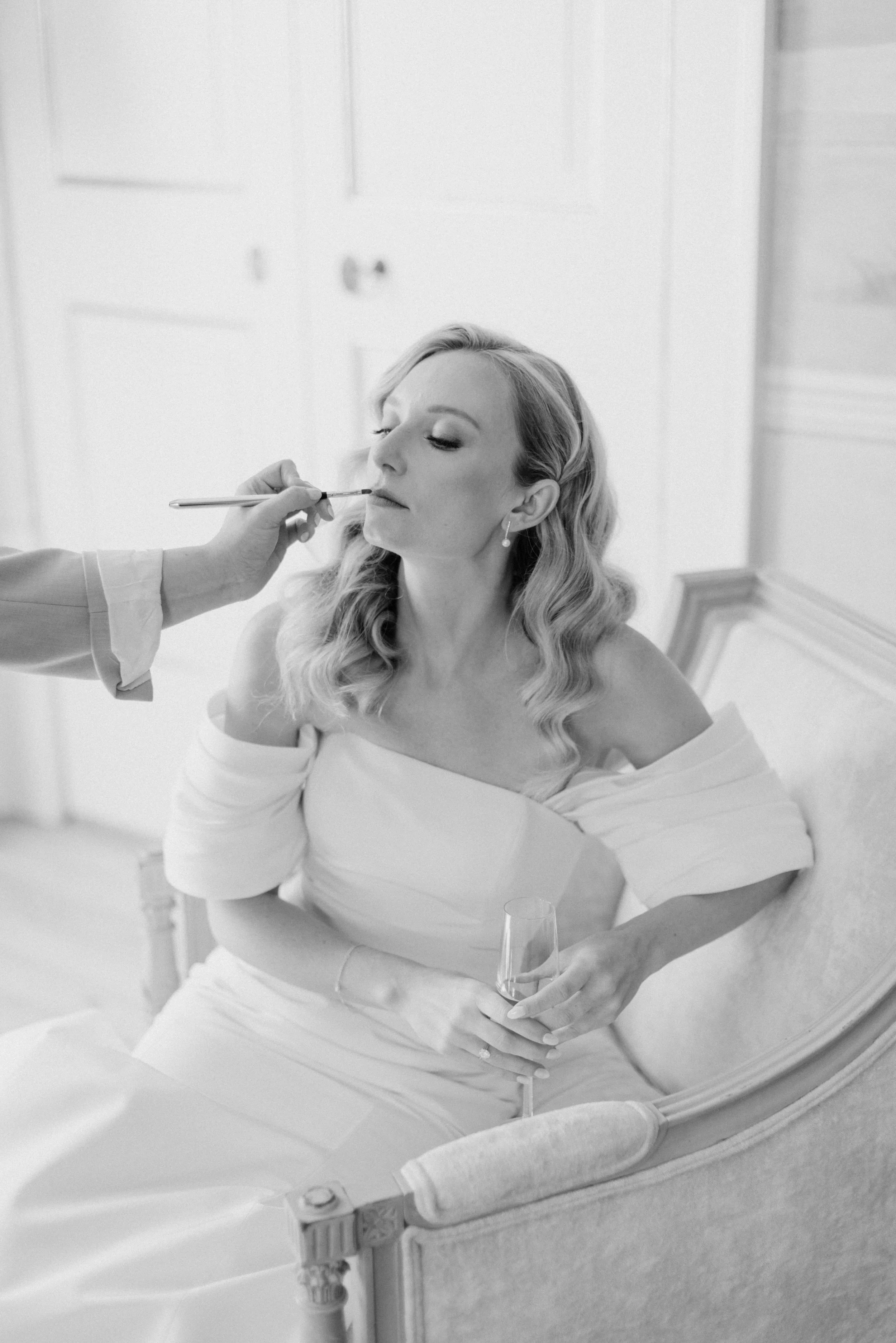A bride sitting in a vanity chair, getting her makeup applied by another person. She is holding a glass of wine and looking slightly upward.