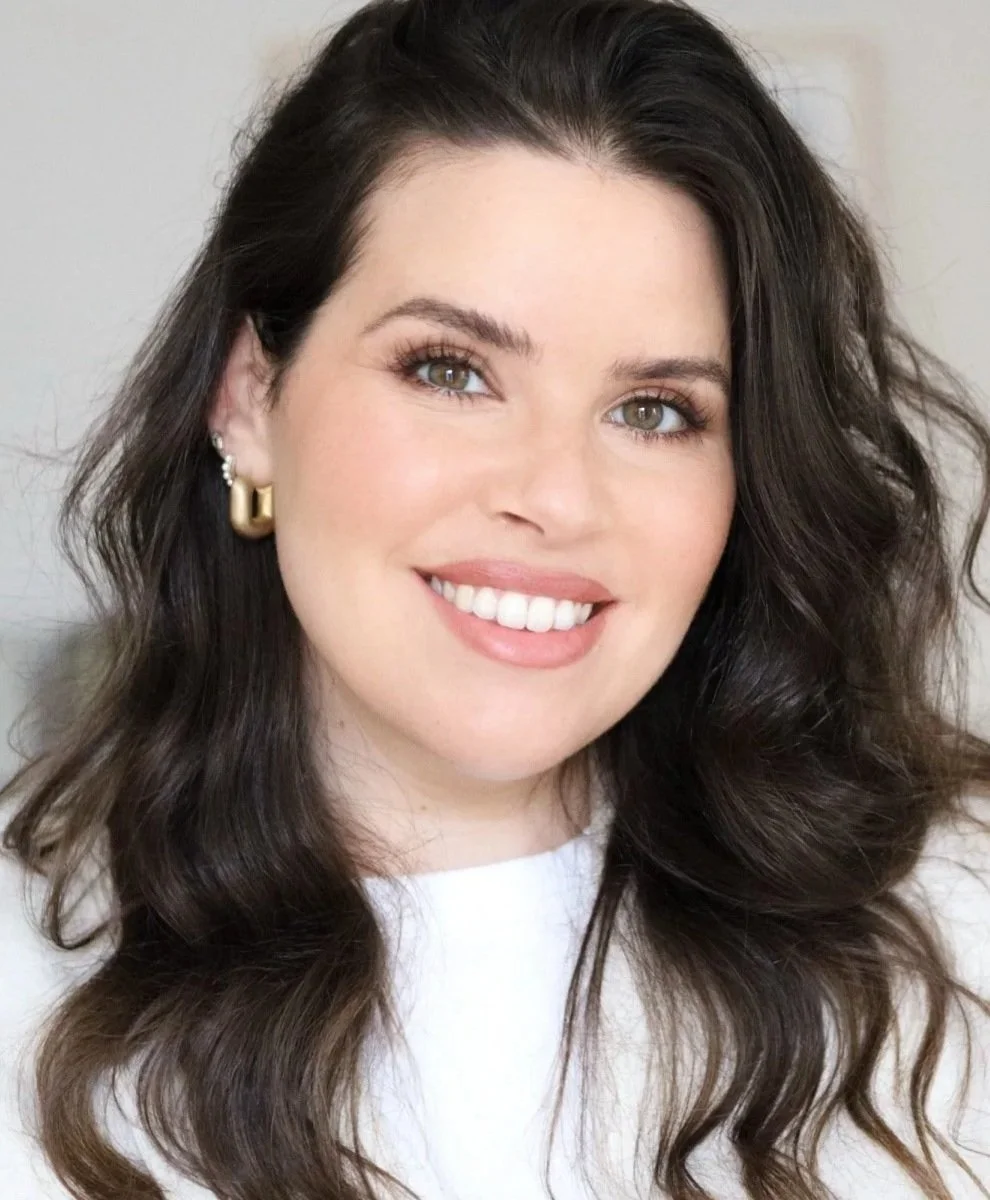 A smiling woman with wavy dark brown hair, wearing a white top and gold earrings.