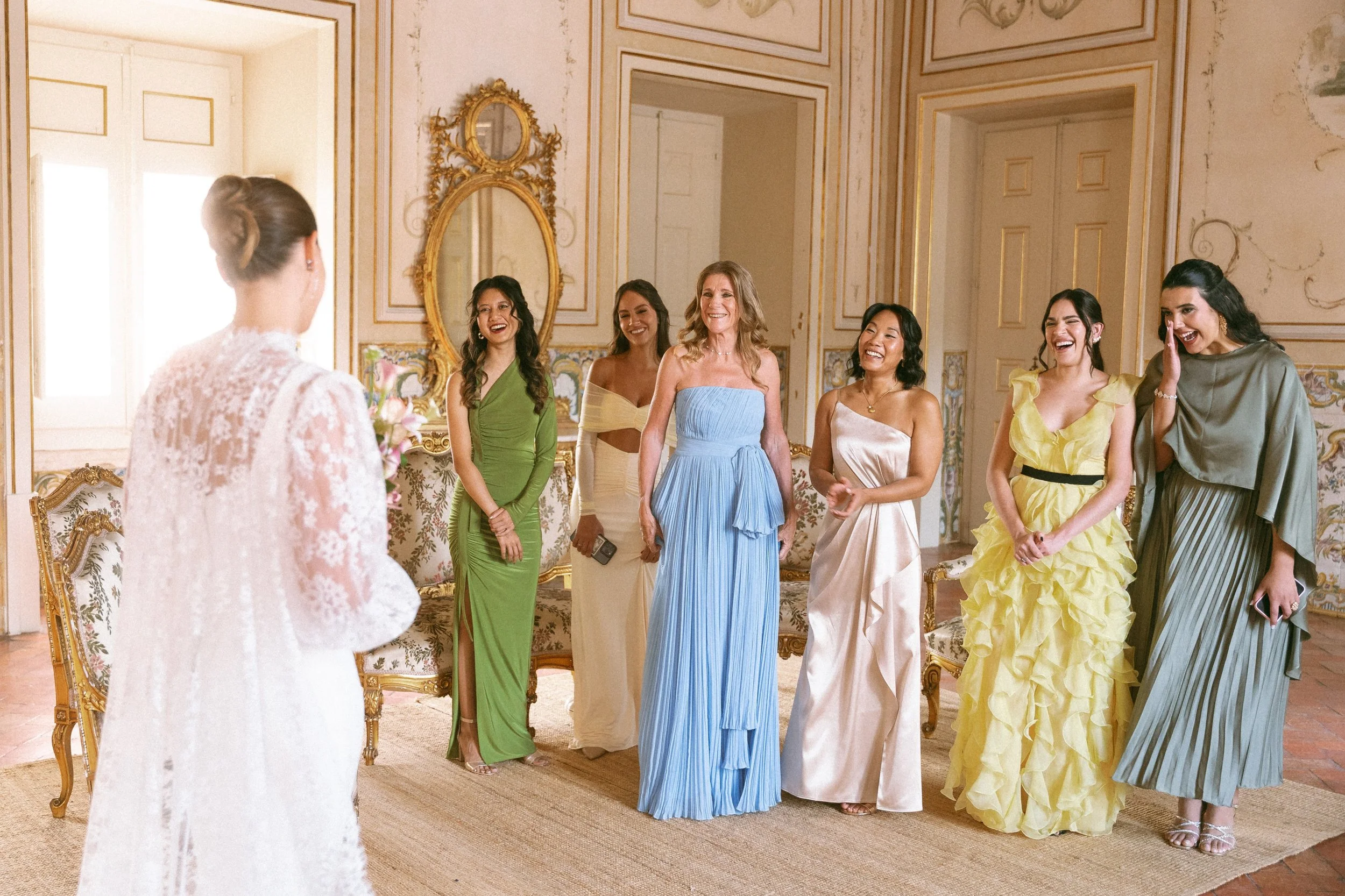 A group of women dressed in colorful evening gowns standing in a luxurious room, smiling and engaging with a woman in a white lace dress who is holding a bouquet of flowers.