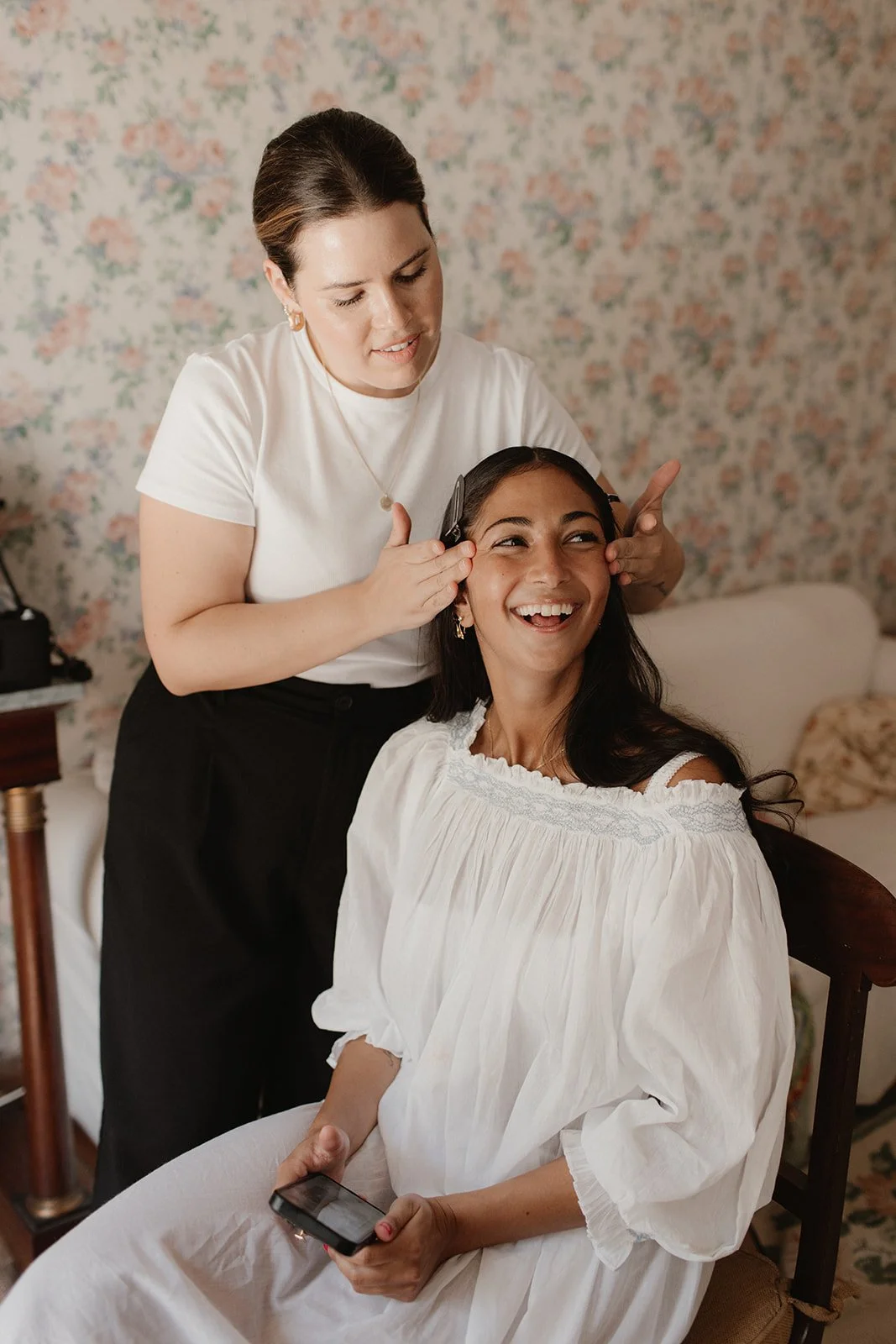A woman with dark hair, wearing a white dress, sitting in a chair and smiling, as a makeup artist styles her hair with a clip, in a room with floral wallpaper.