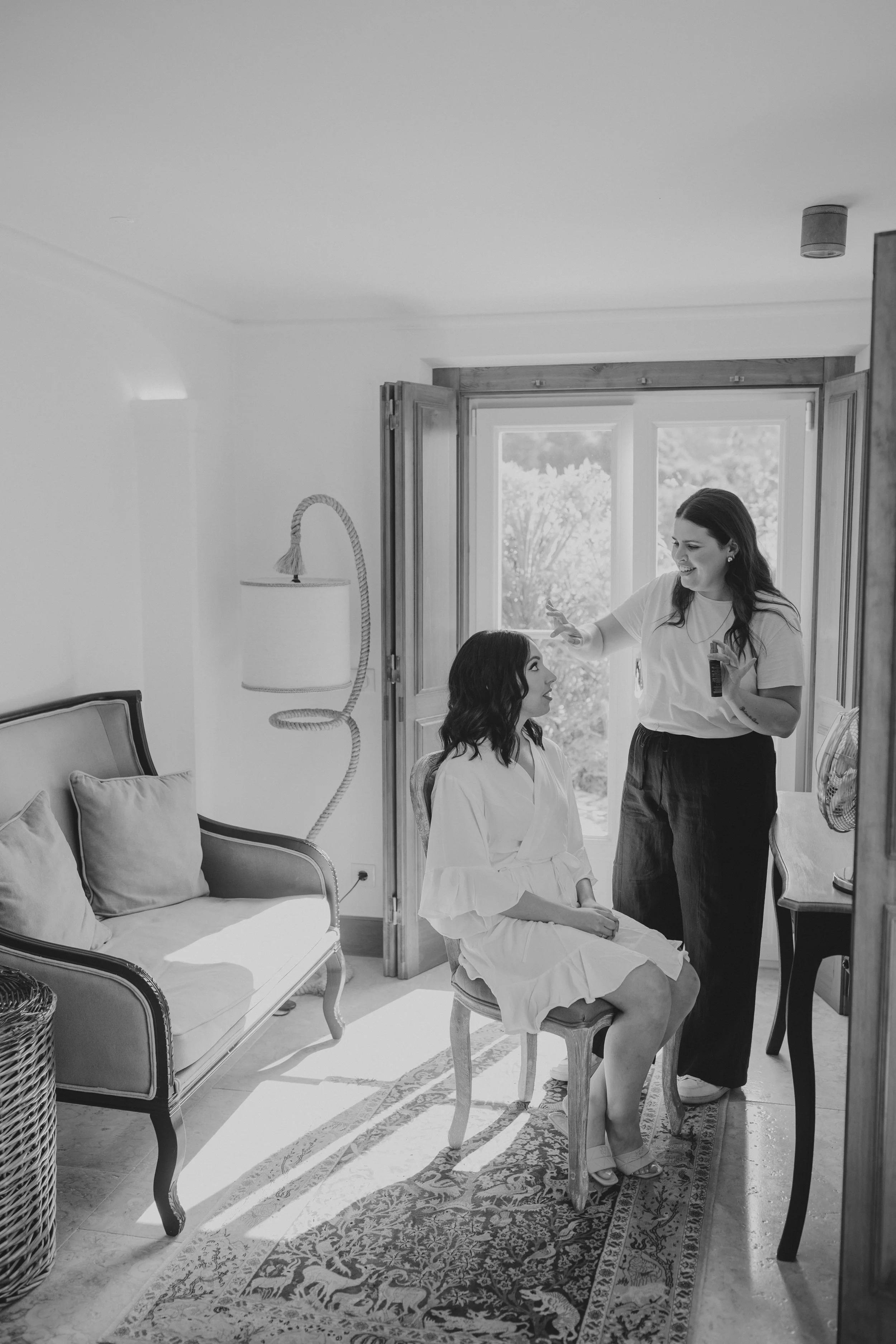 A bride in a white robe sitting on a chair, getting her makeup done by another woman standing next to her in a bright room with open doorway and sunlight.