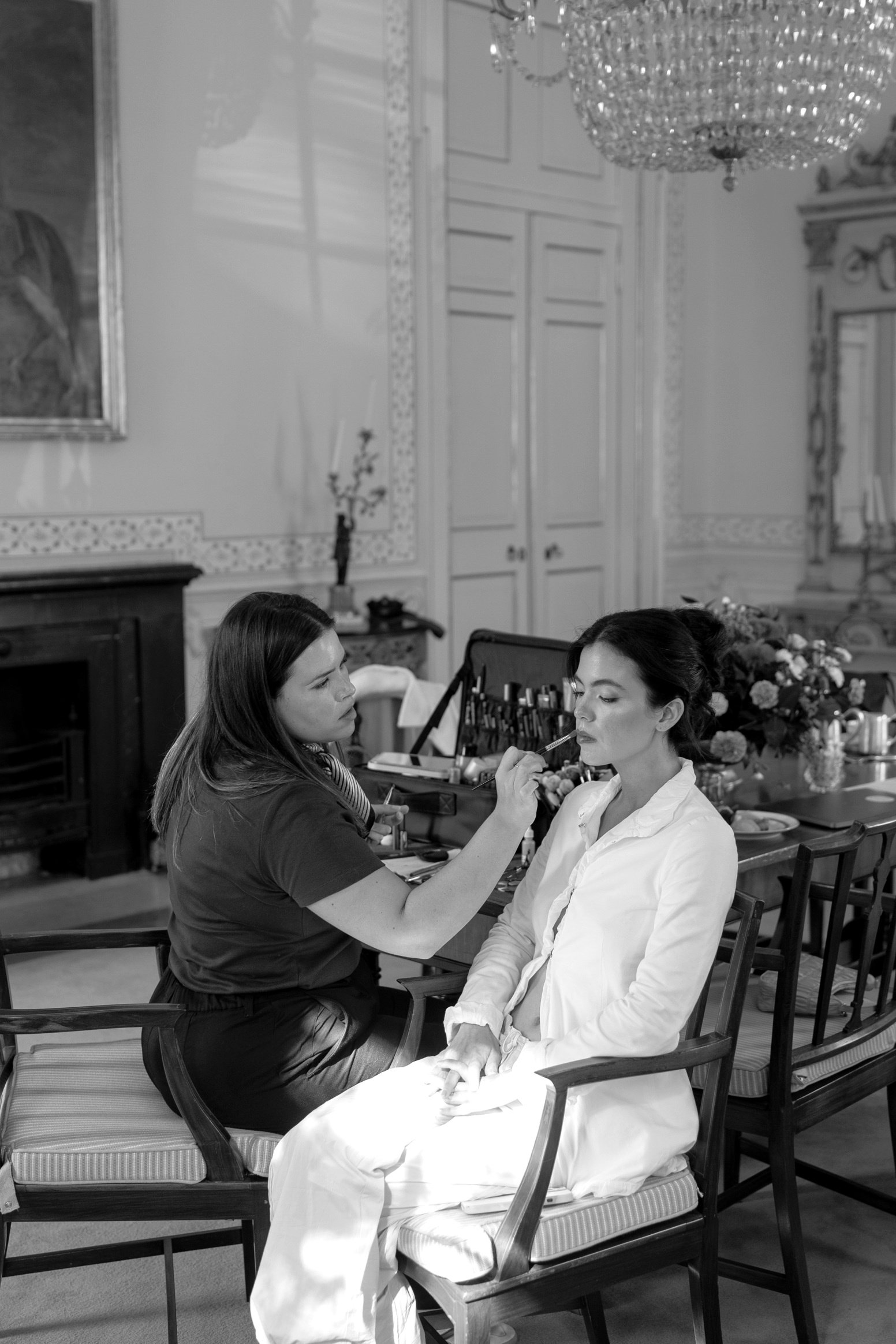 Woman in a wedding venue getting their makeup done, with a makeup artist applying lipstick to a seated woman in a formal room.