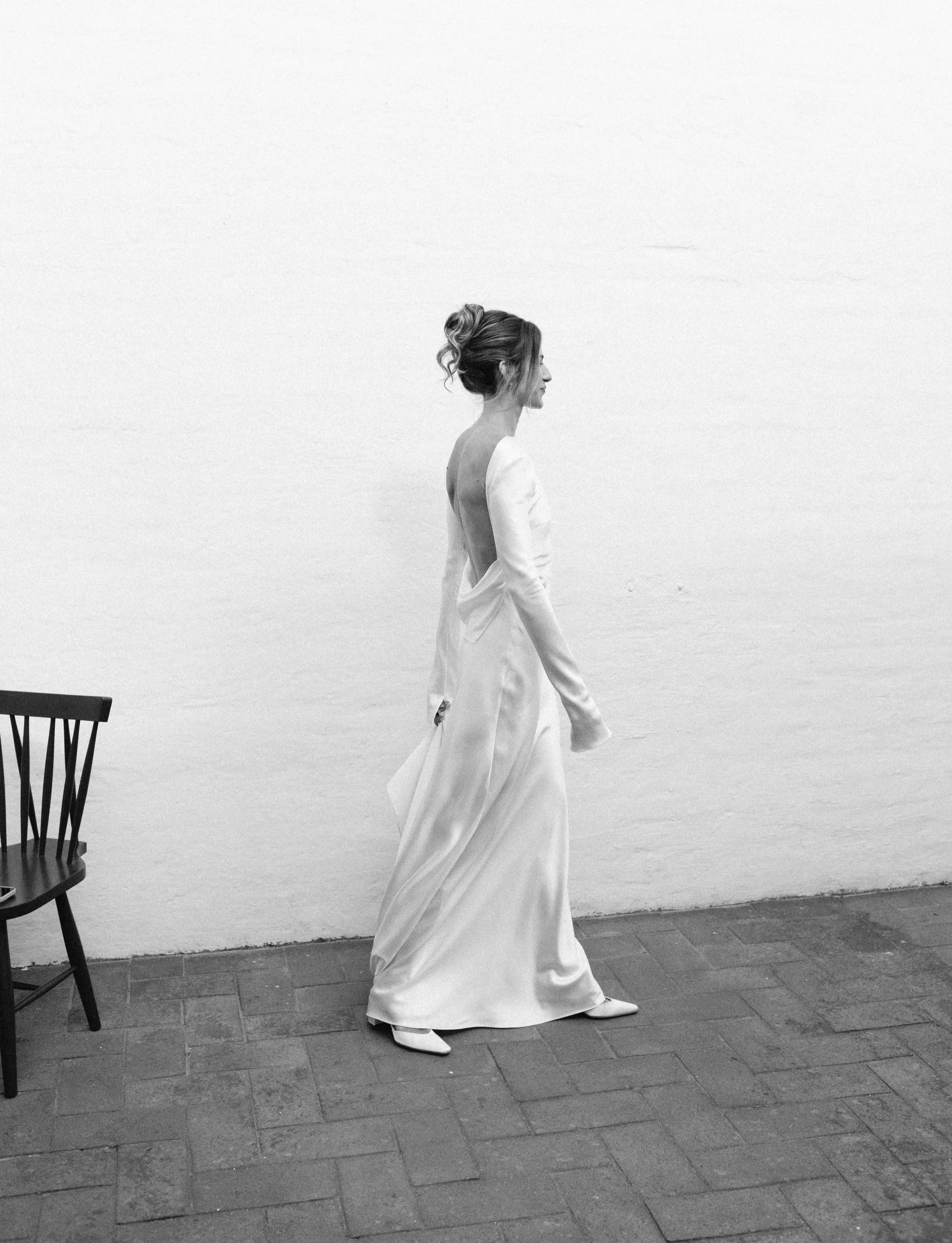 Black and white photo of a woman in a white dress standing against a plain wall, with a chair nearby.