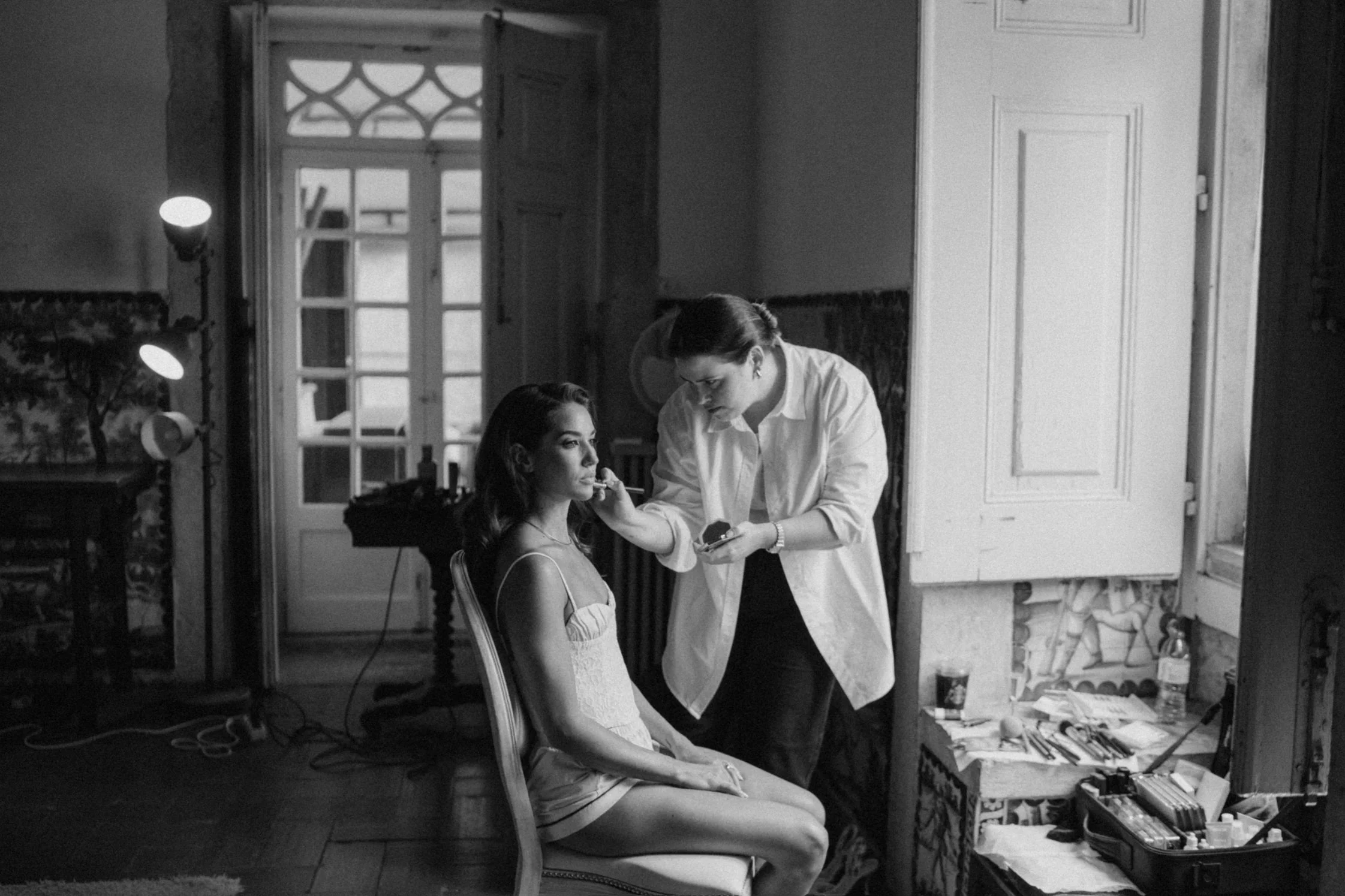 A woman sitting on a chair while another woman applies makeup to her face in a room with a window, a door, and a table with makeup supplies.