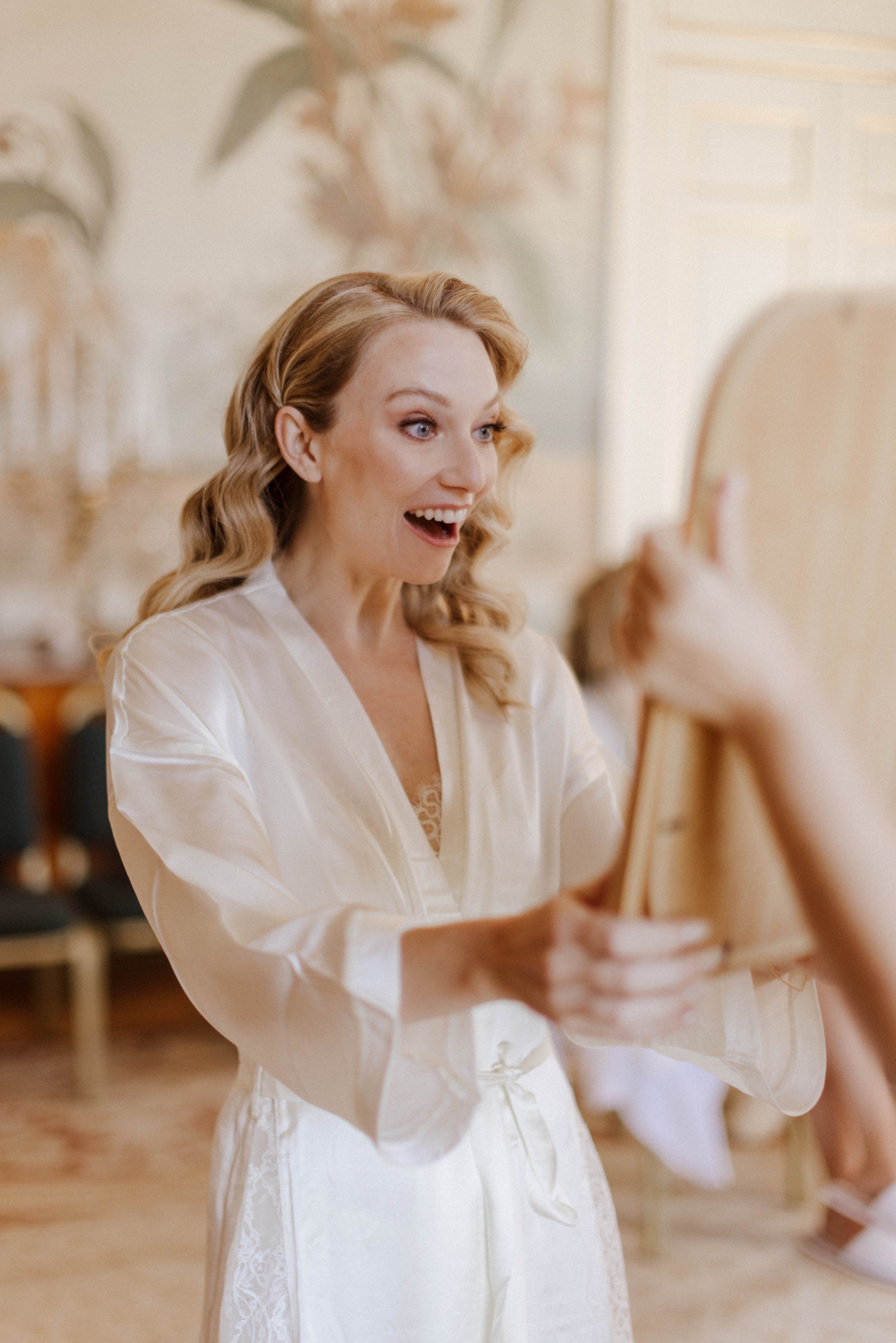 A woman with blonde, wavy hair smiling while looking at herself in a mirror, wearing a white satin robe with lace details, in a warmly lit room with abstract artwork on the wall behind her.