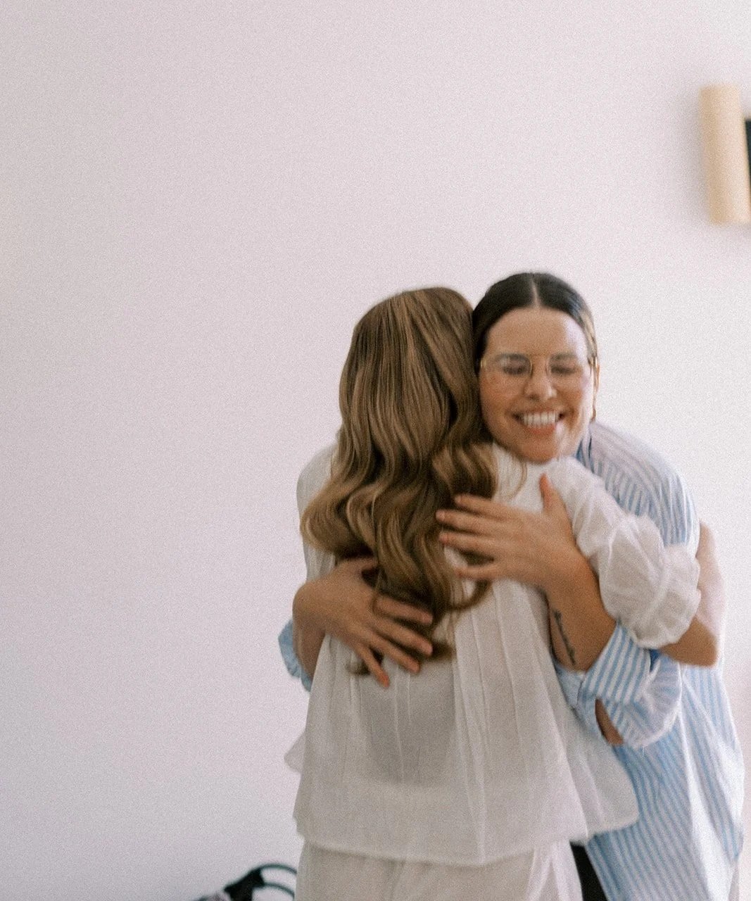 Two women hugging, one with wavy brown hair and the other with straight black hair, smiling with eyes closed against a plain wall.